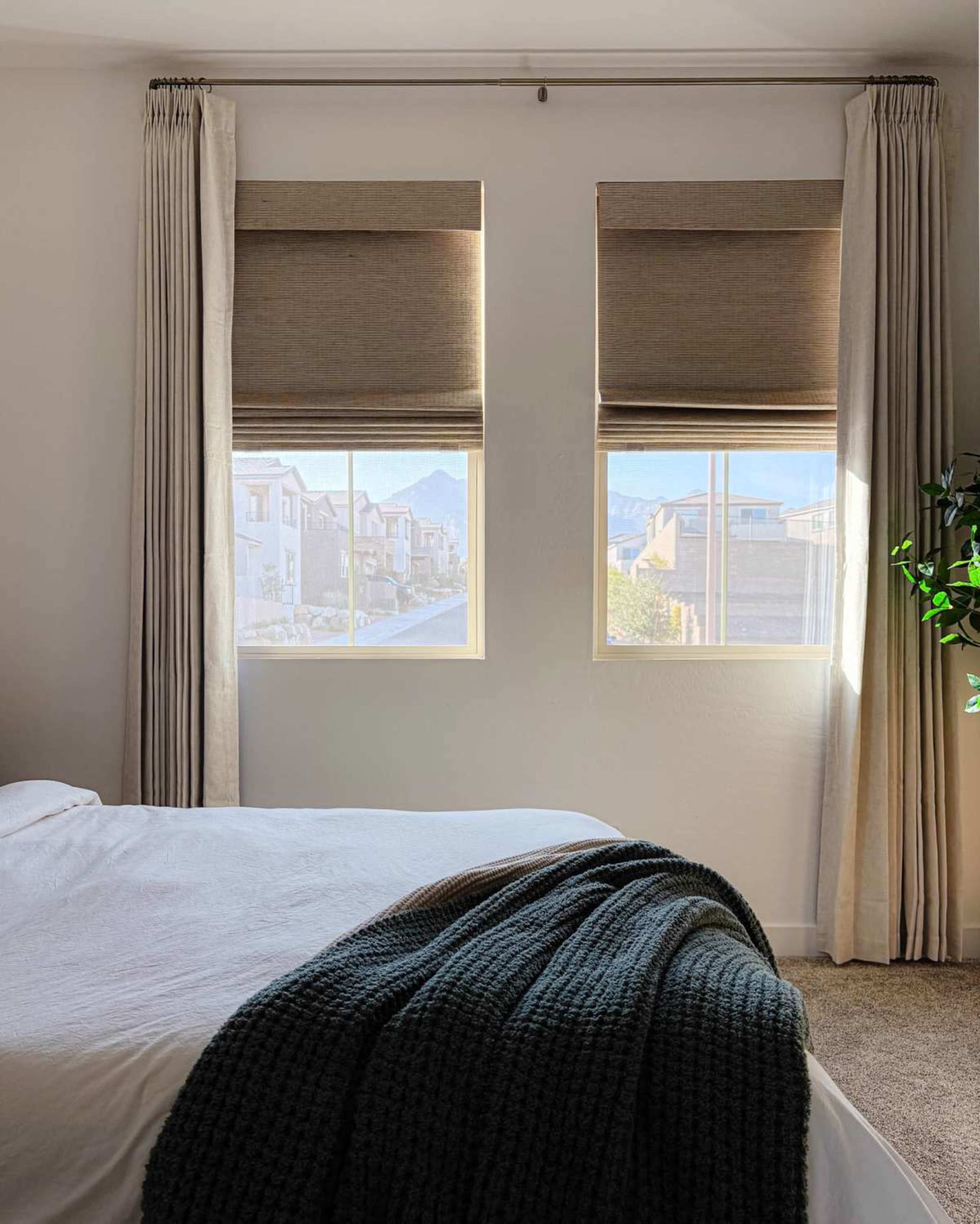 A bedroom features a bed with a white blanket, two windows with shades, and curtains, allowing natural light to illuminate the space.