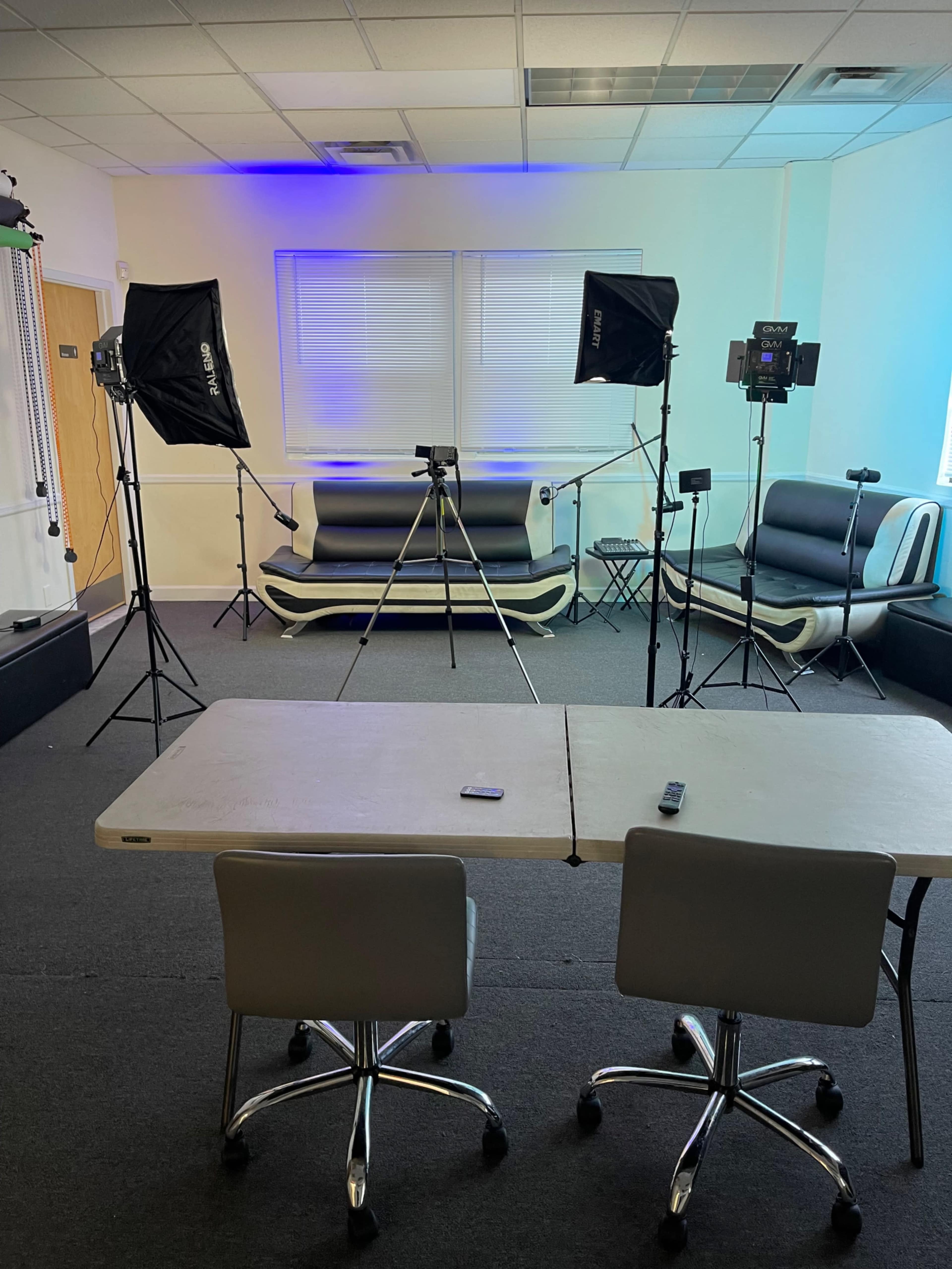 The image shows a studio setup with lighting equipment, two folding chairs, and several black leather couches arranged in a room with gray carpet.