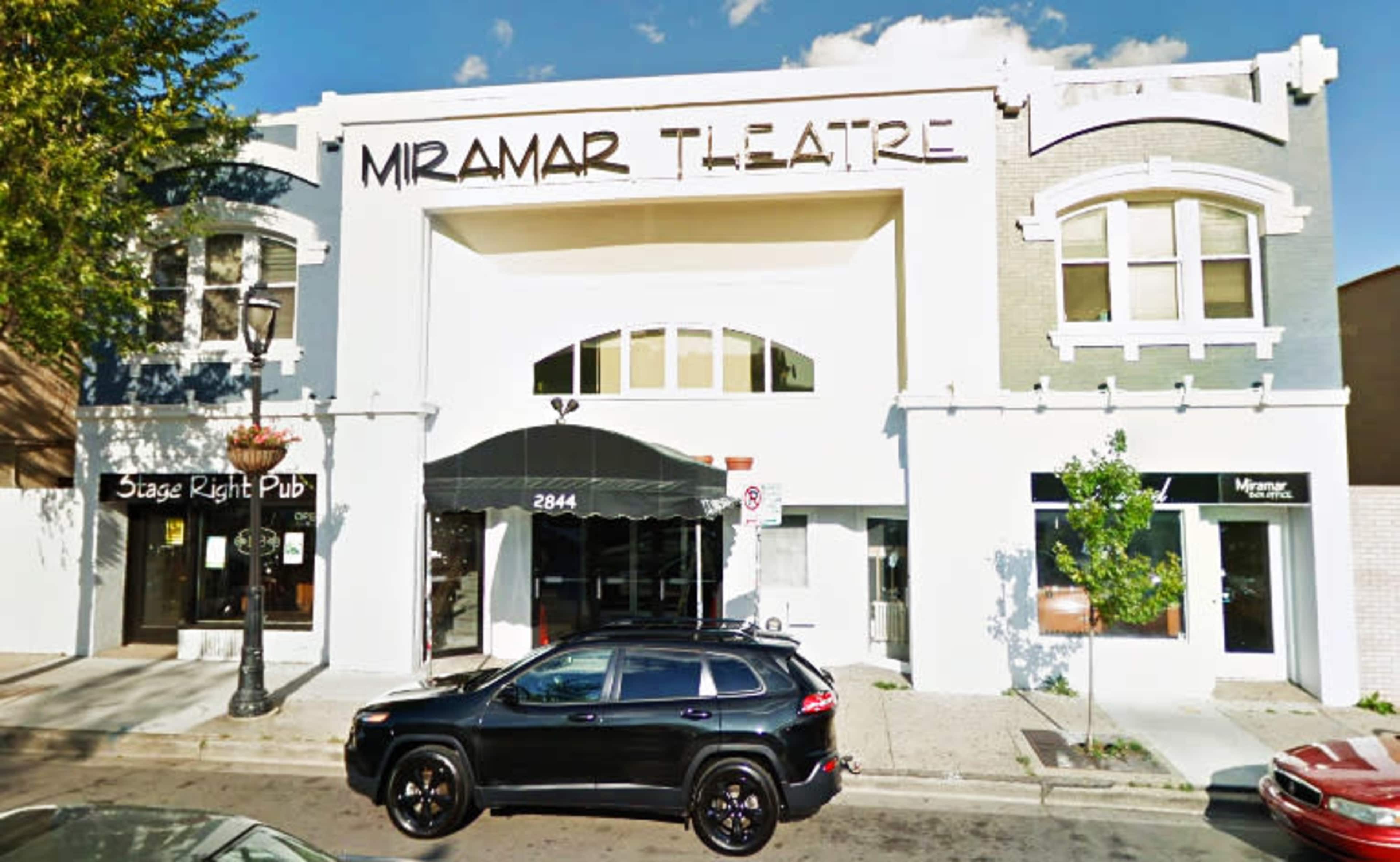 The image shows the Miramar Theatre building with a black SUV parked in front and a clear blue sky.