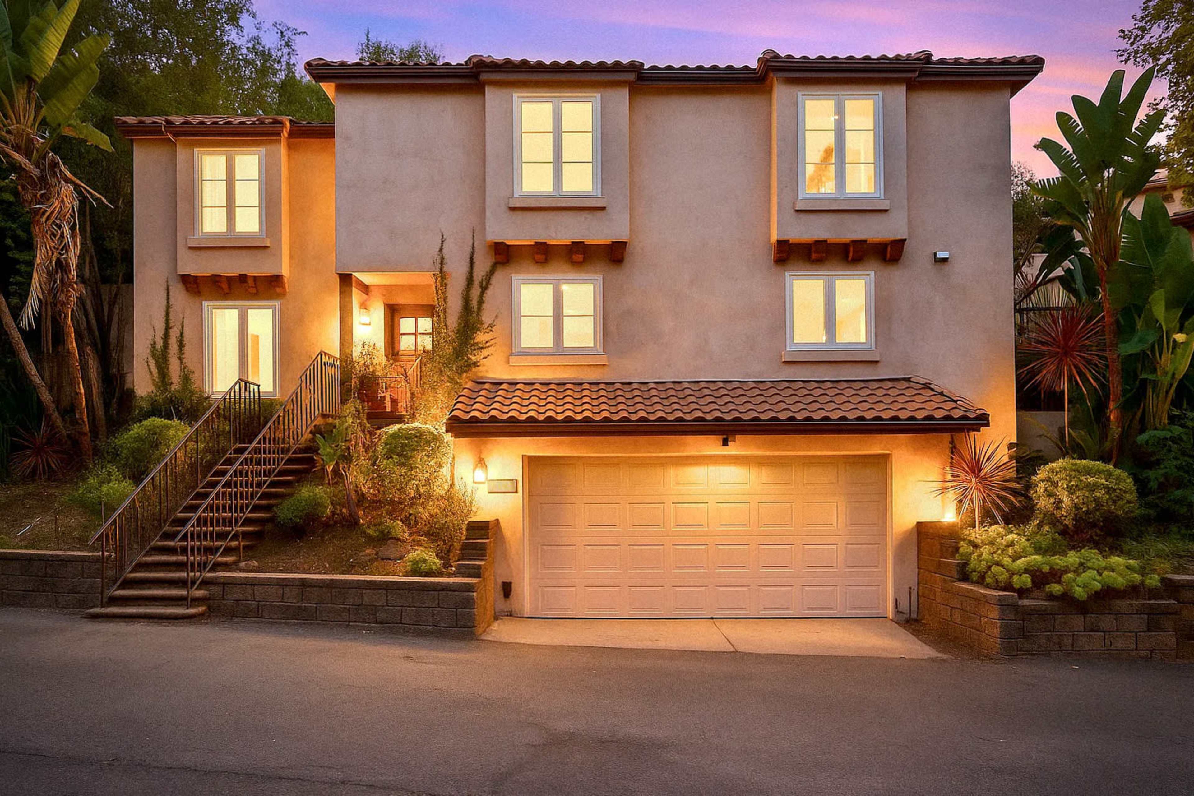The image shows a two-story house with a tile roof, featuring a garage and a landscaped front yard, illuminated at dusk.