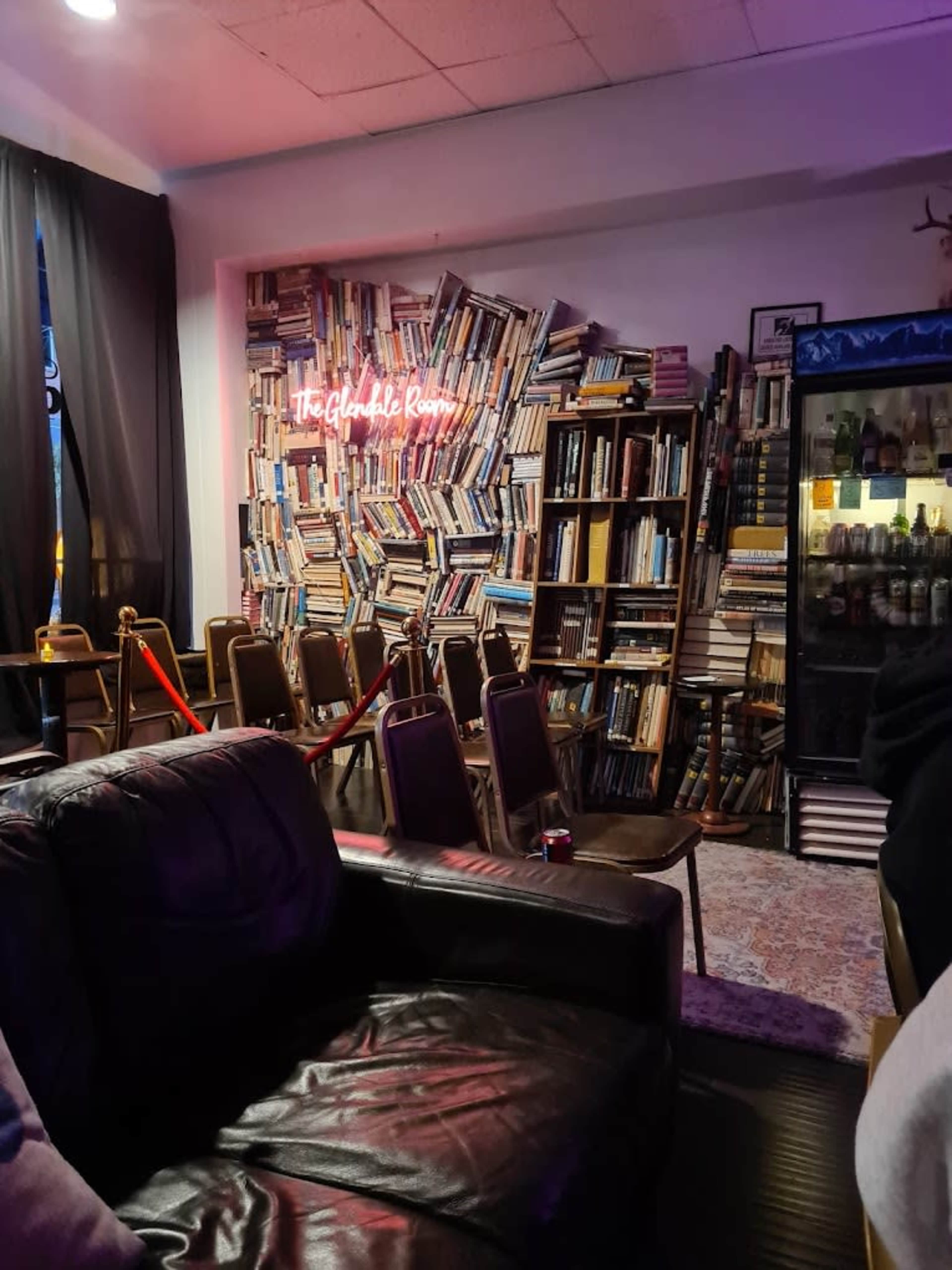 A cozy room features a wall of stacked books, a neon sign reading "The Glanville Room," and a row of chairs set up in front of a display case.