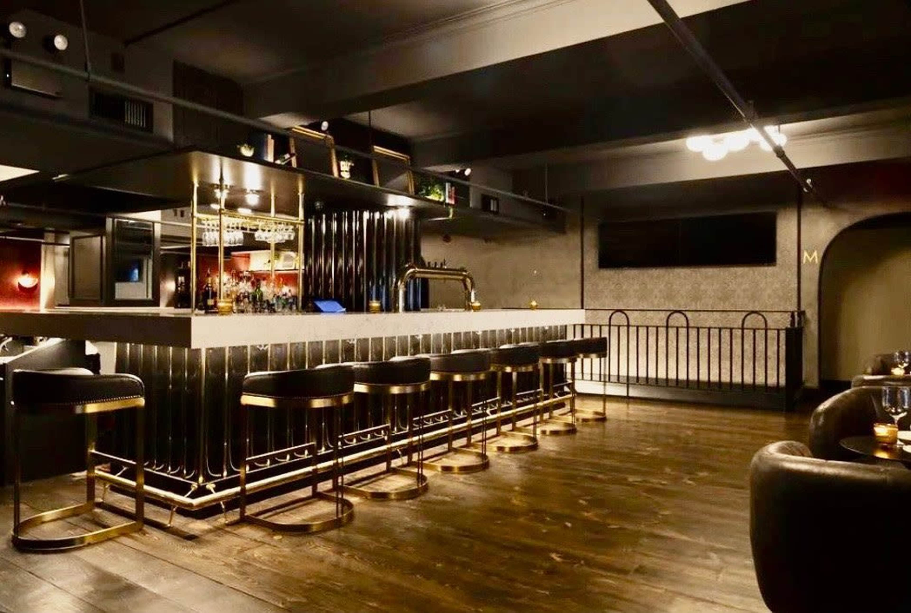 The image shows a modern bar with a sleek design, featuring a long countertop, high stools, and a variety of liquor displayed on shelves.