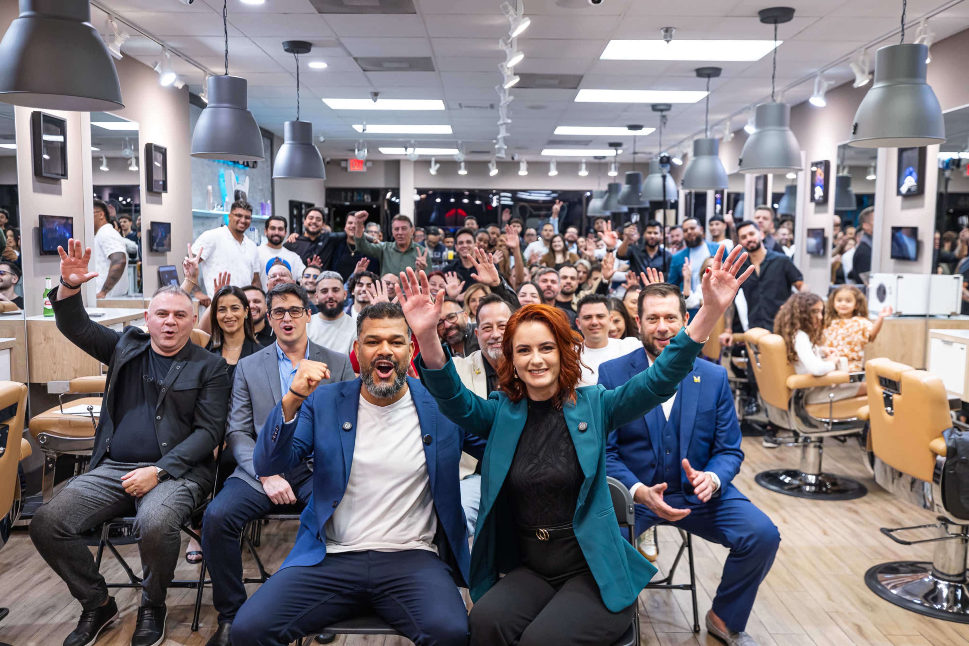 A large group of people is gathered in a barbershop, with some individuals seated and others standing, all appearing to celebrate and cheer towards the camera.