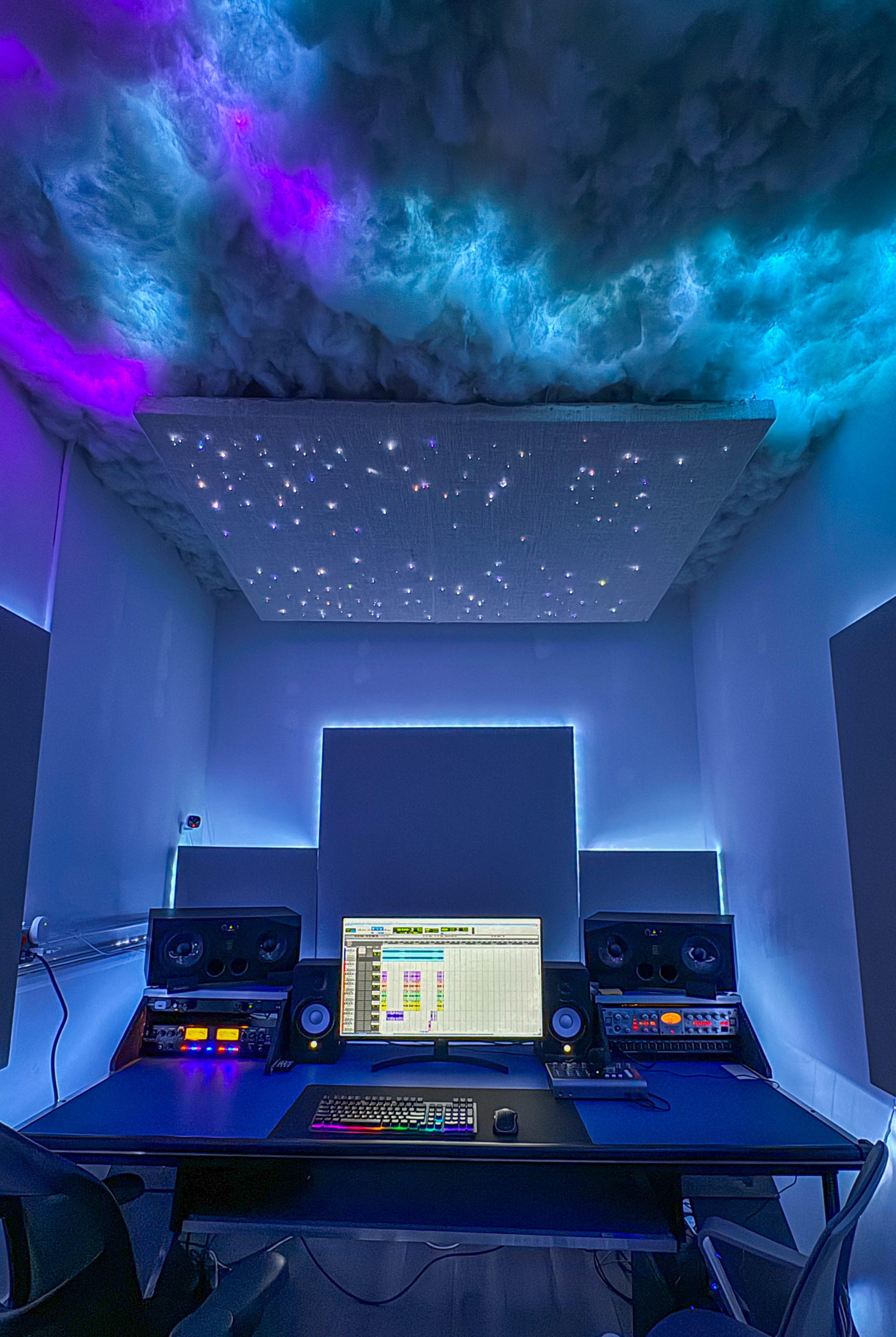 A modern music studio features a dark desk with audio equipment and a ceiling illuminated with colorful lighting effects.