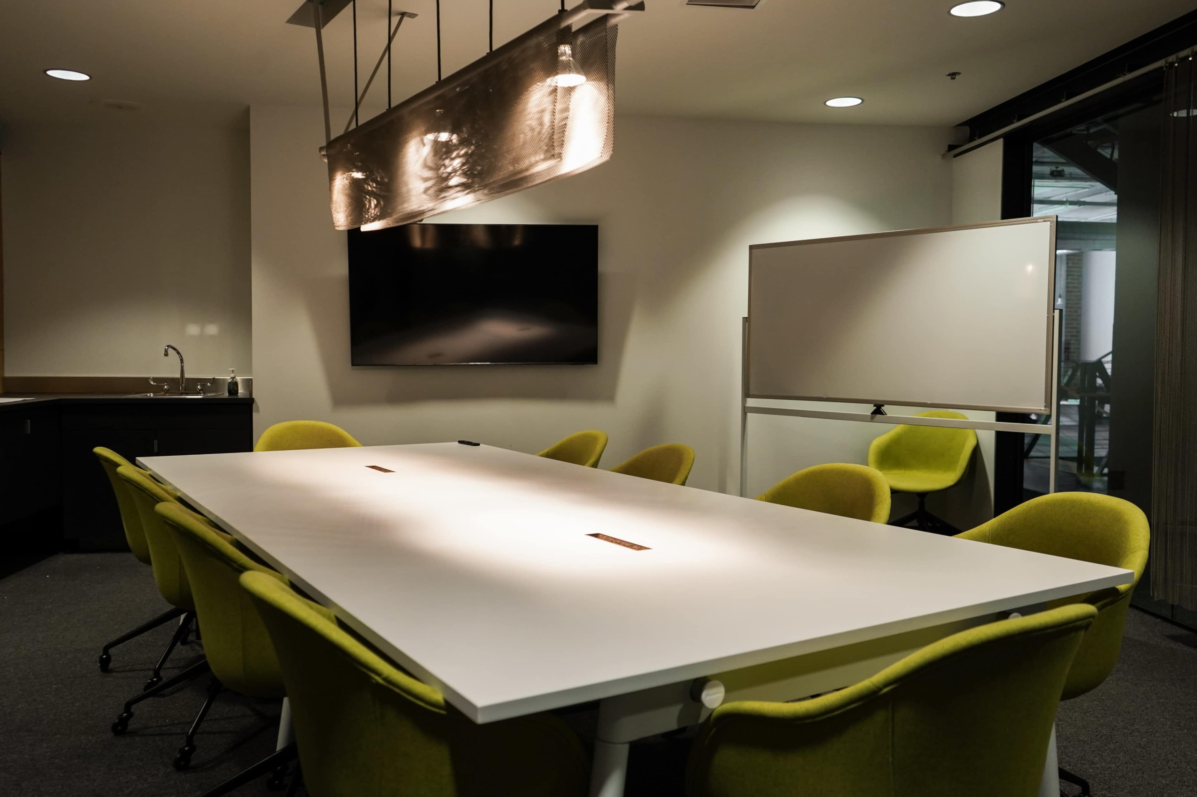 The image shows a modern conference room featuring a long white table surrounded by yellow chairs, a digital screen on the wall, and a whiteboard.