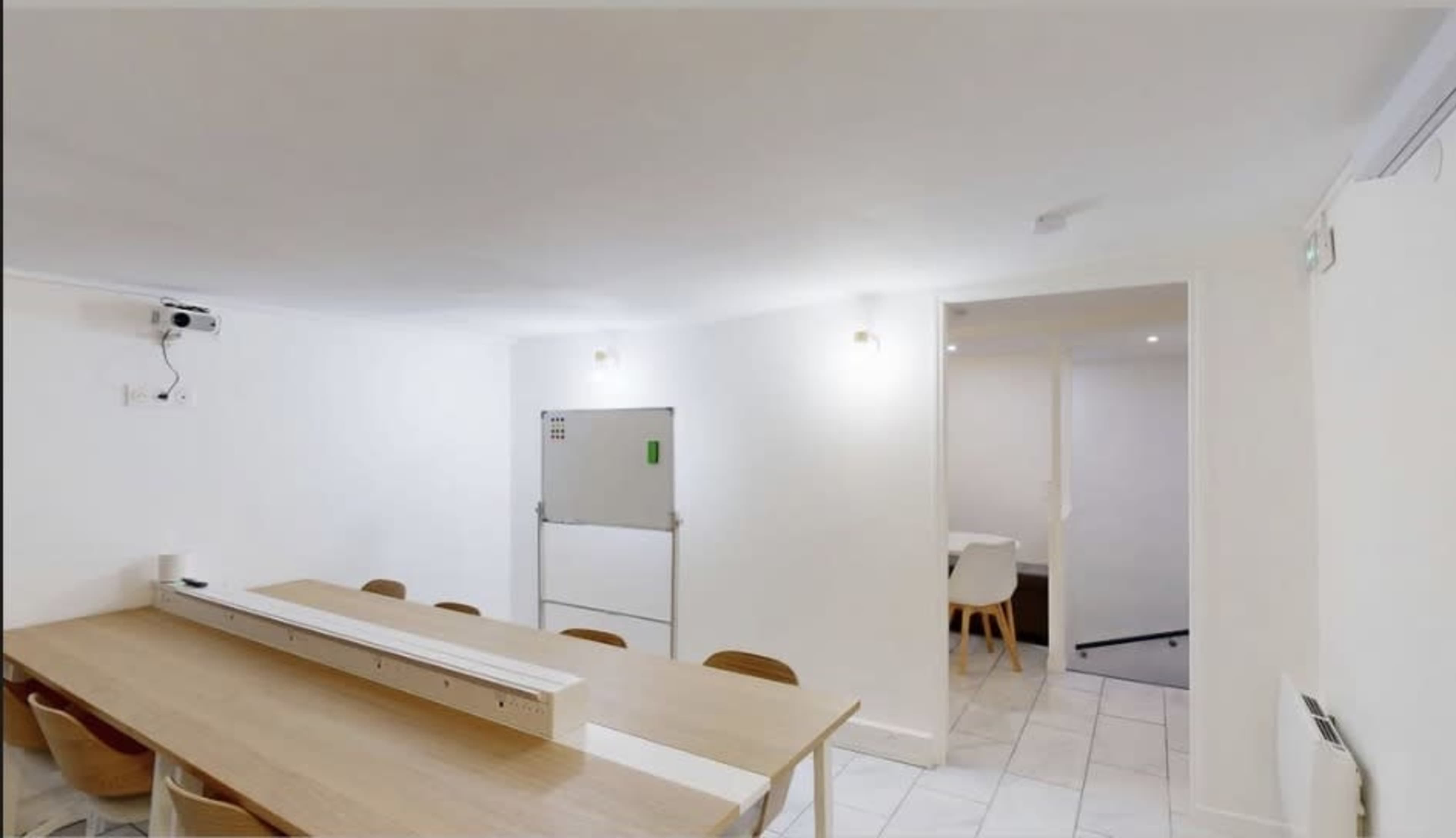 The image shows a well-lit, minimalist meeting room with a long wooden table and chairs, featuring a projection screen on one wall and a doorway leading to another space.