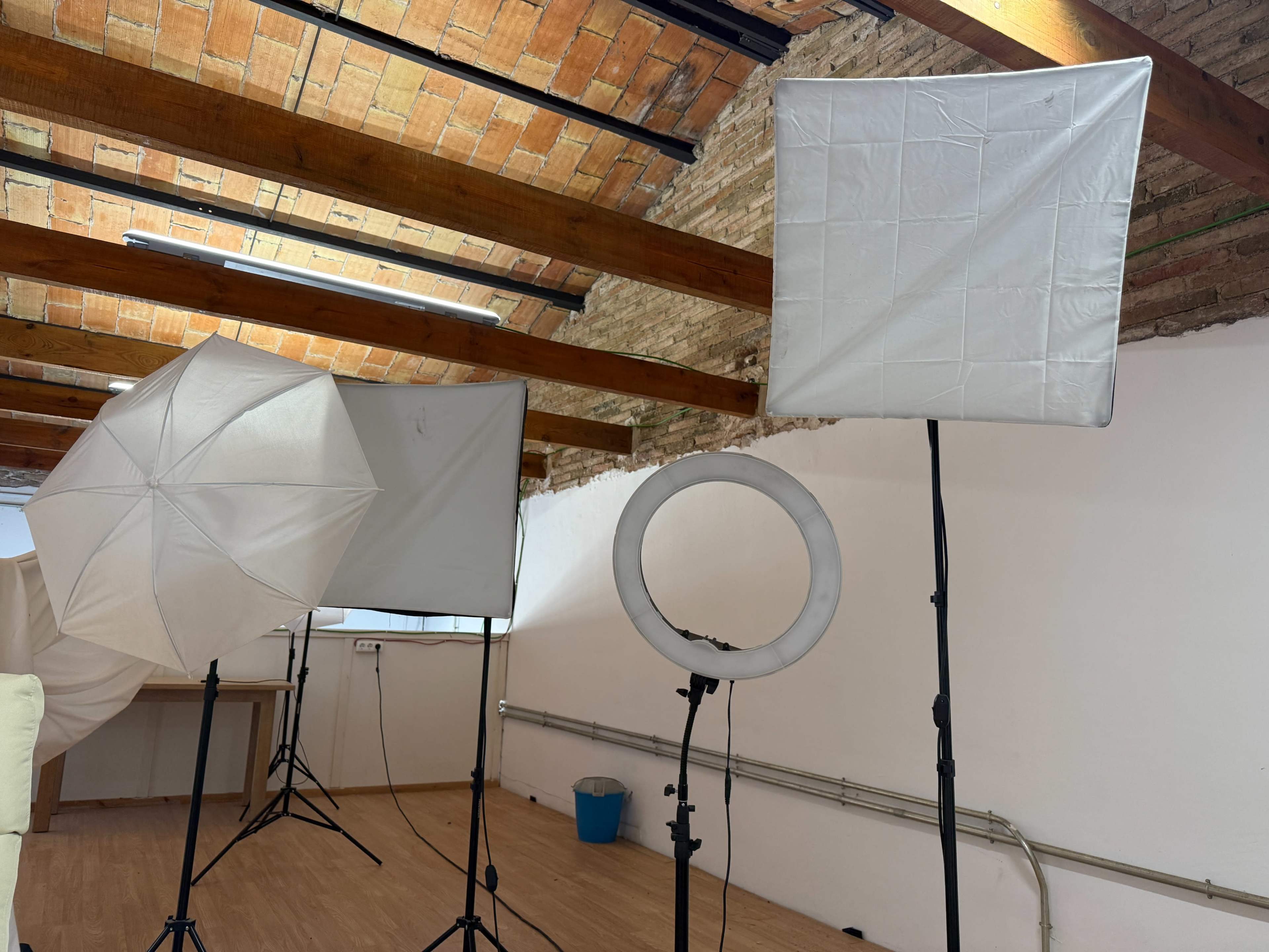 The scene shows various photography lighting equipment, including softboxes and a ring light, set up in a room with exposed brick walls and wooden beams.