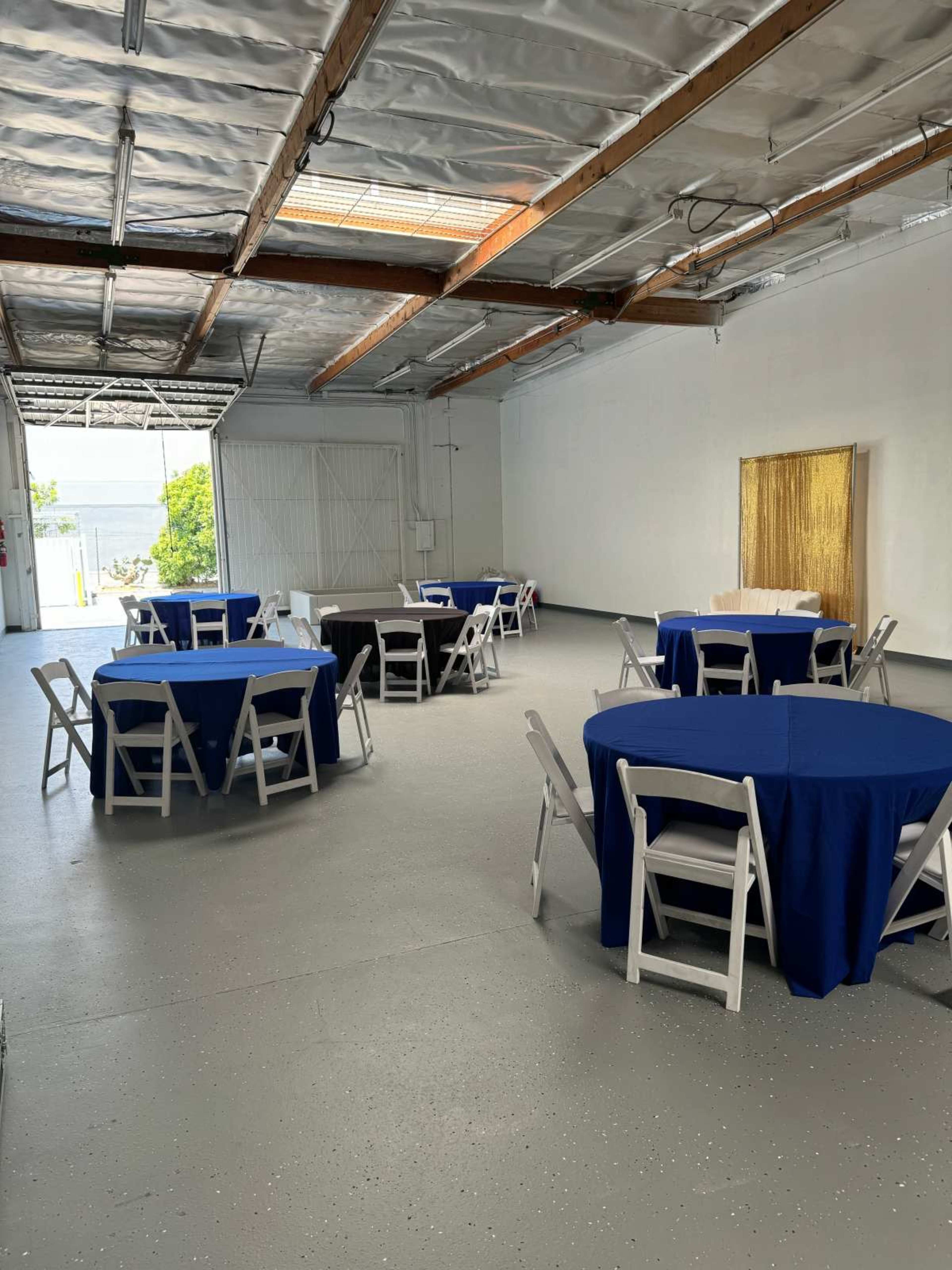 A spacious room features several round tables covered with blue and black tablecloths, arranged in a clean and open layout.