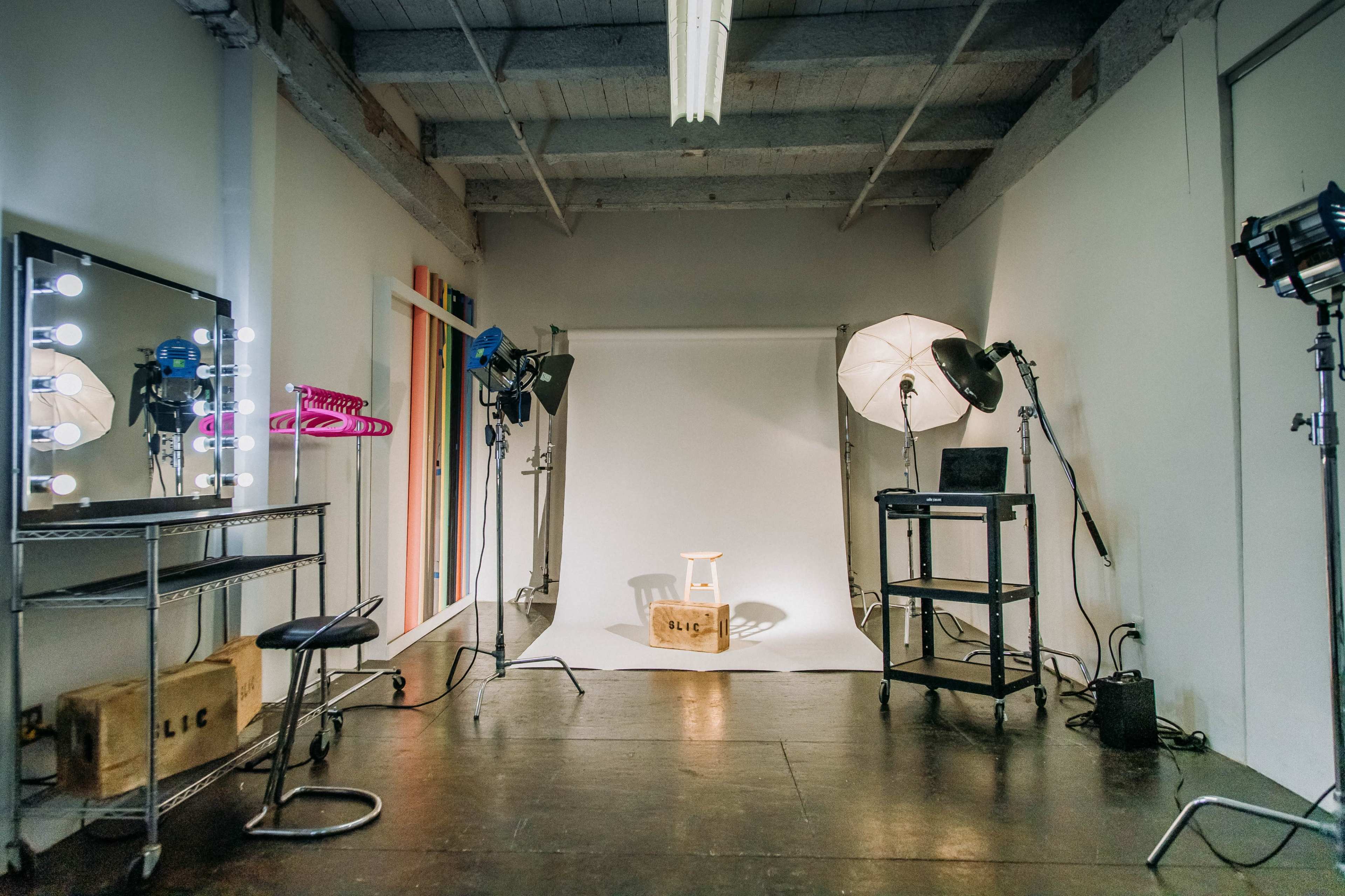 A photography studio features a light backdrop, a chair on a stand, and various lighting equipment arranged around the space.