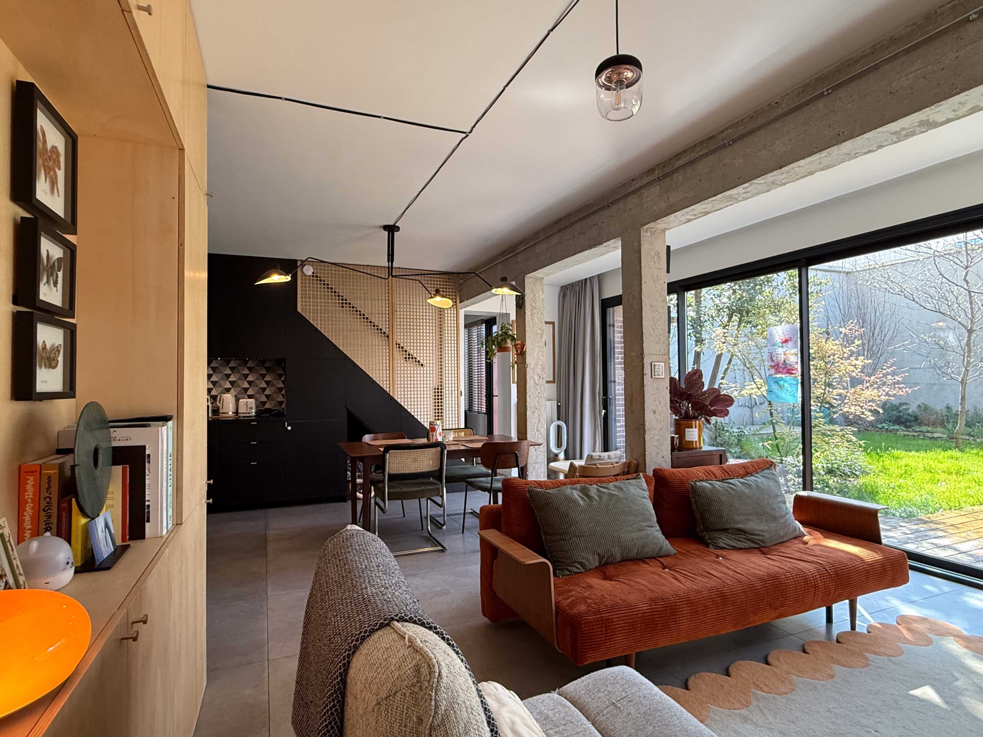 The image shows a modern living space with a textured orange sofa, a dining area in the background, and large glass doors leading to an outdoor garden.