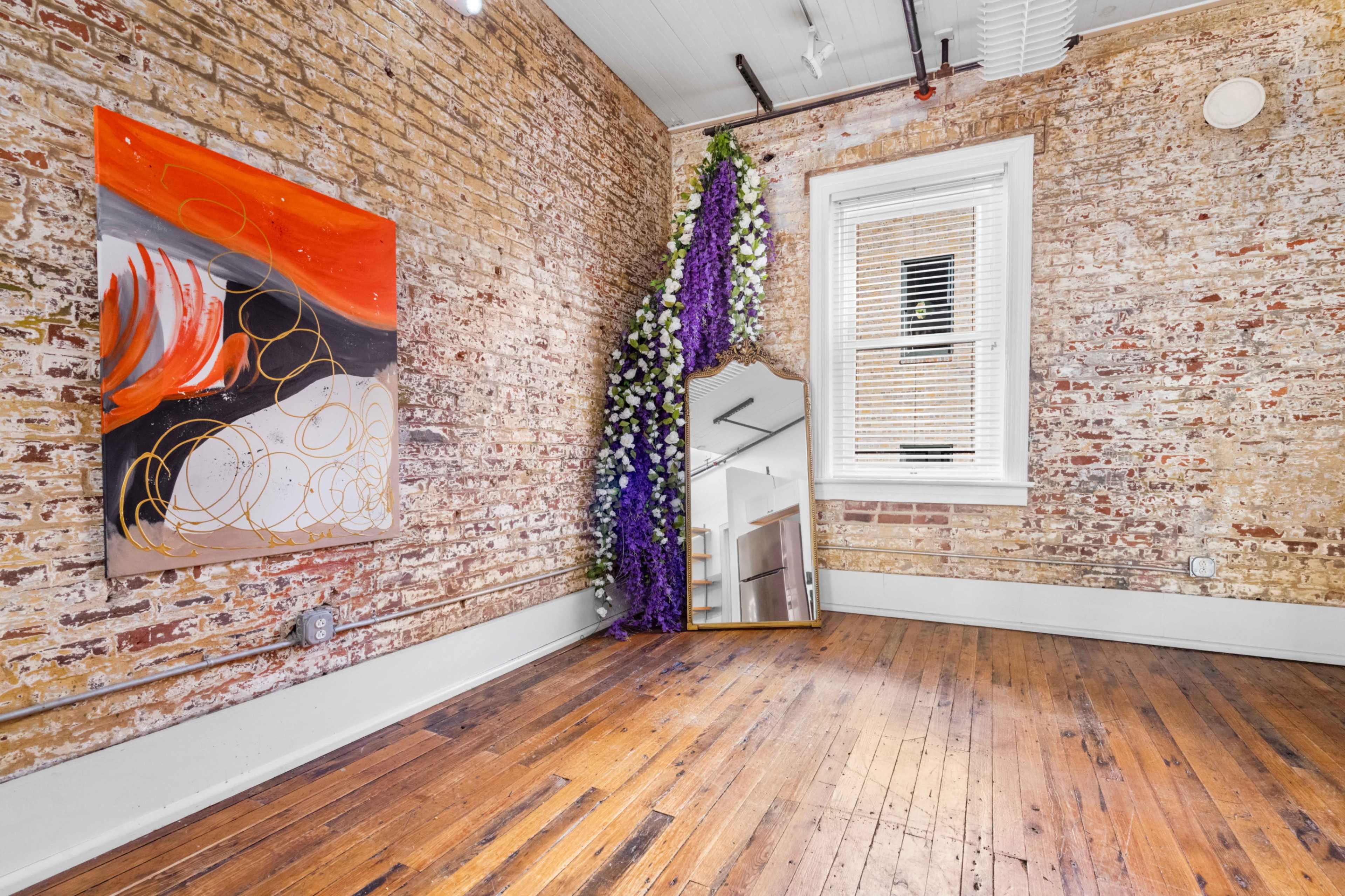 The image shows a room with exposed brick walls, a large mirror, a colorful abstract painting, and artificial flowers draping in one corner.