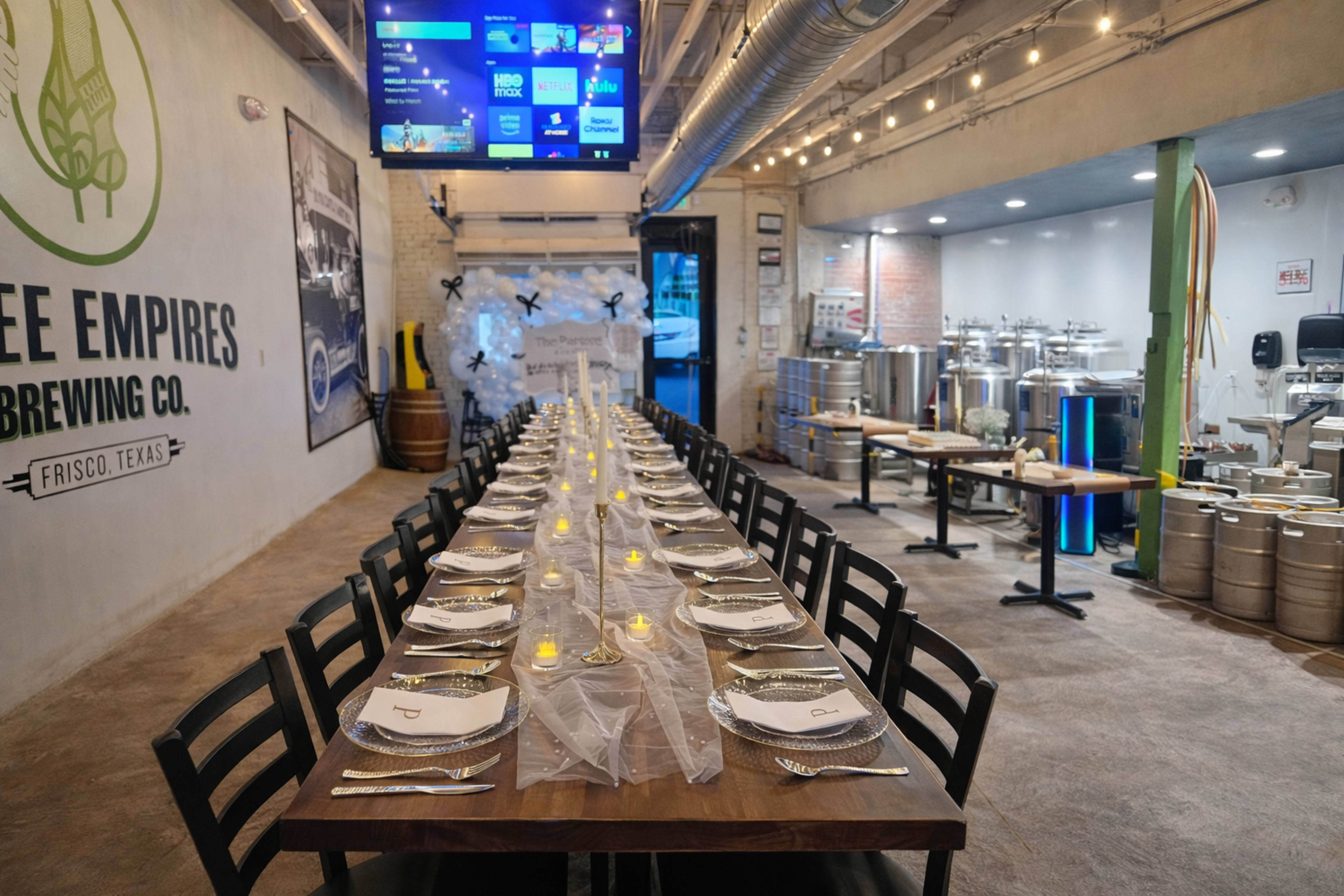 A long, elegantly set dining table with lit candles is positioned in a brewery, with brewing equipment and a large screen visible in the background.