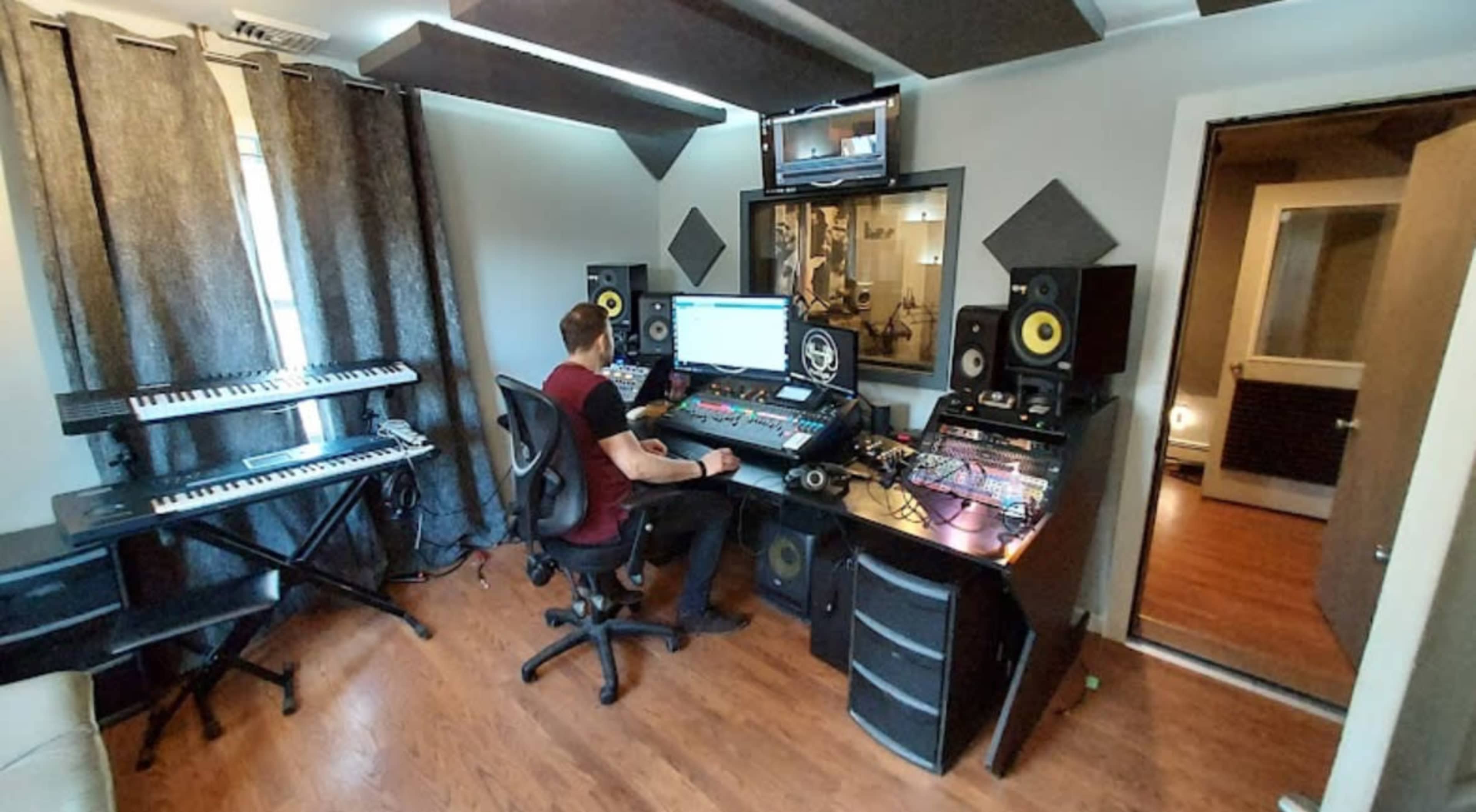 A person is seated at a music production workstation surrounded by various audio equipment and instruments.