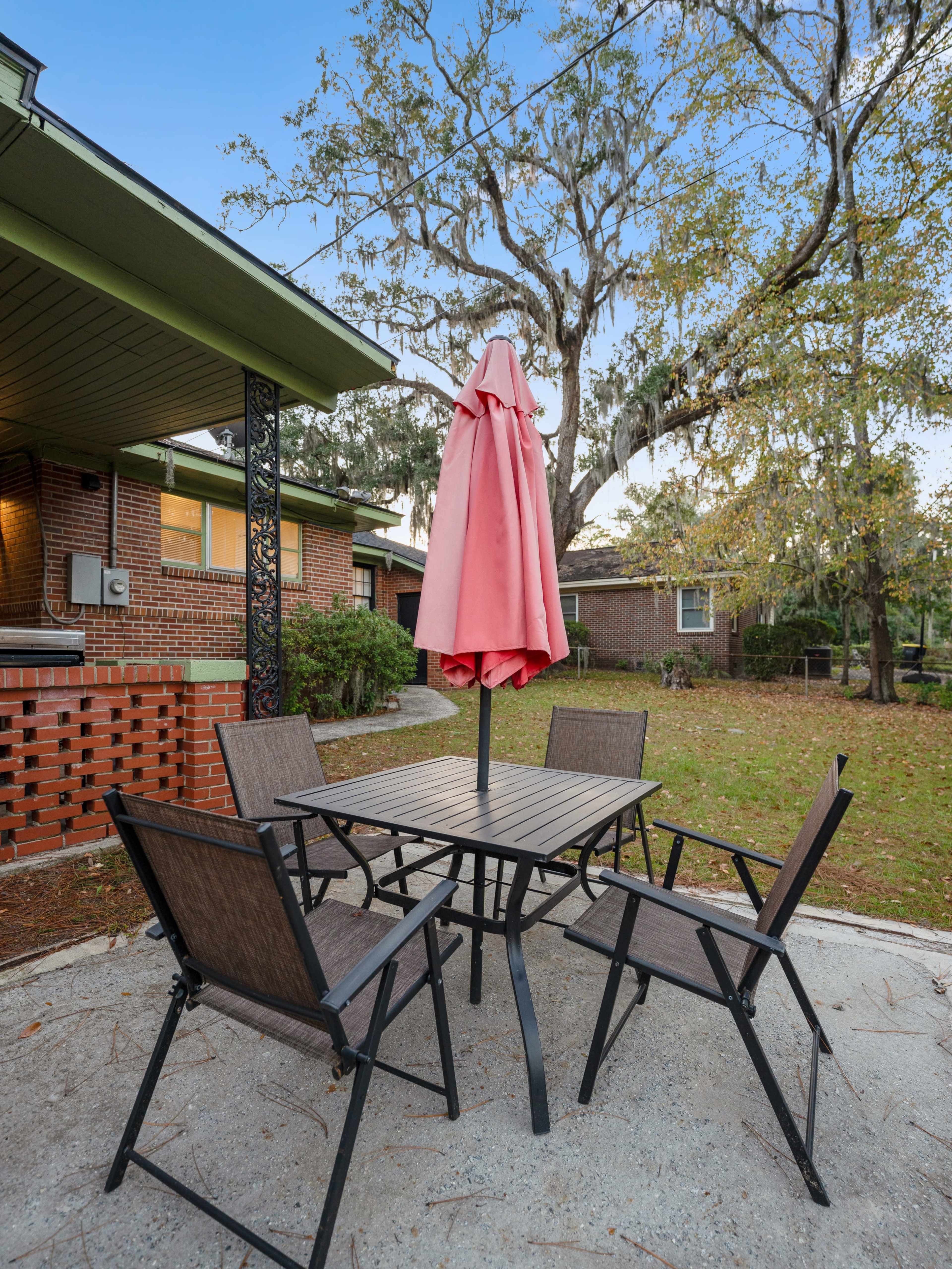A patio table with four chairs and a pink umbrella is situated on a concrete surface in a yard surrounded by trees.