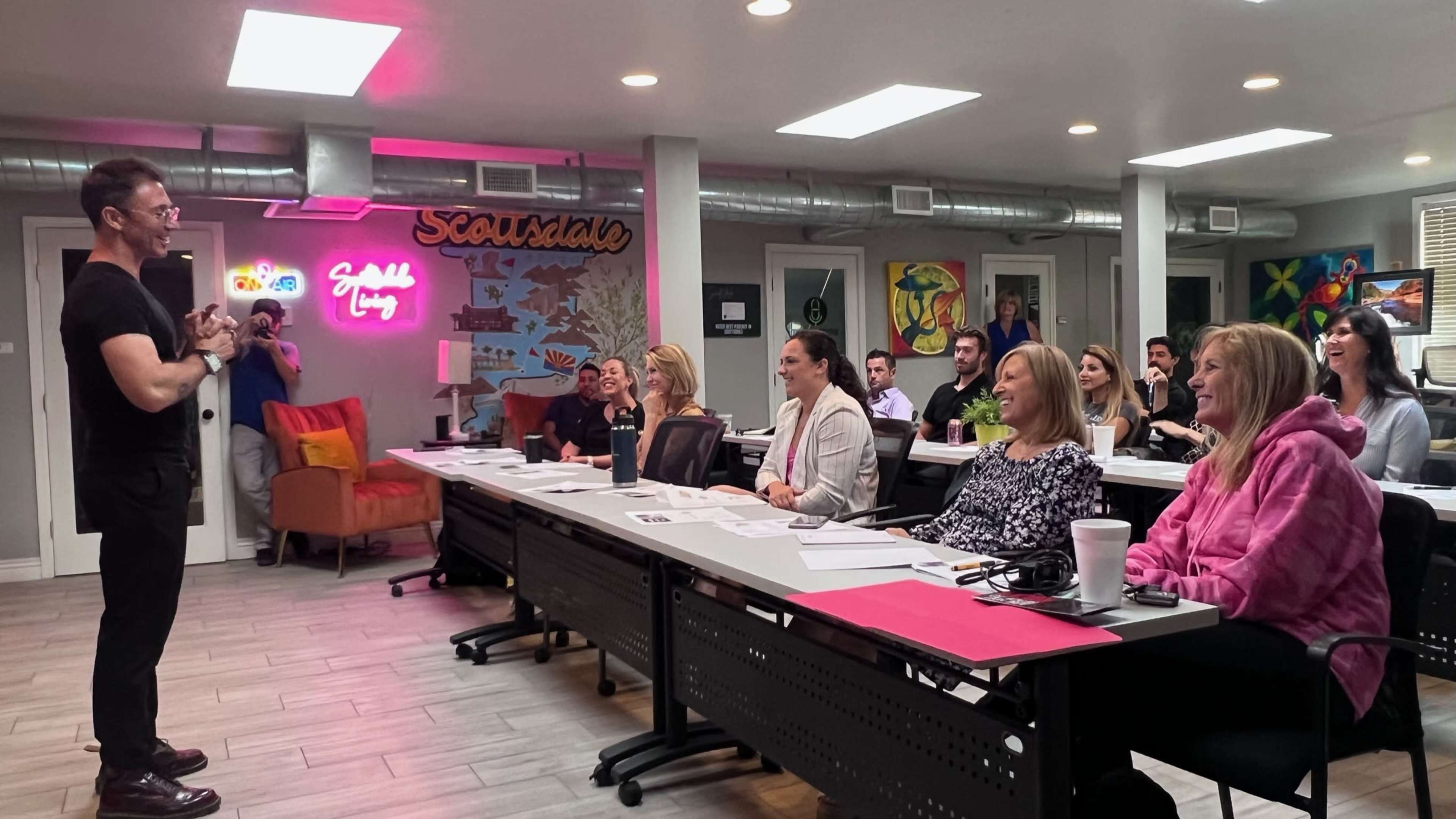 A presenter stands in front of an engaged audience in a modern classroom setting with colorful decor and a neon sign.