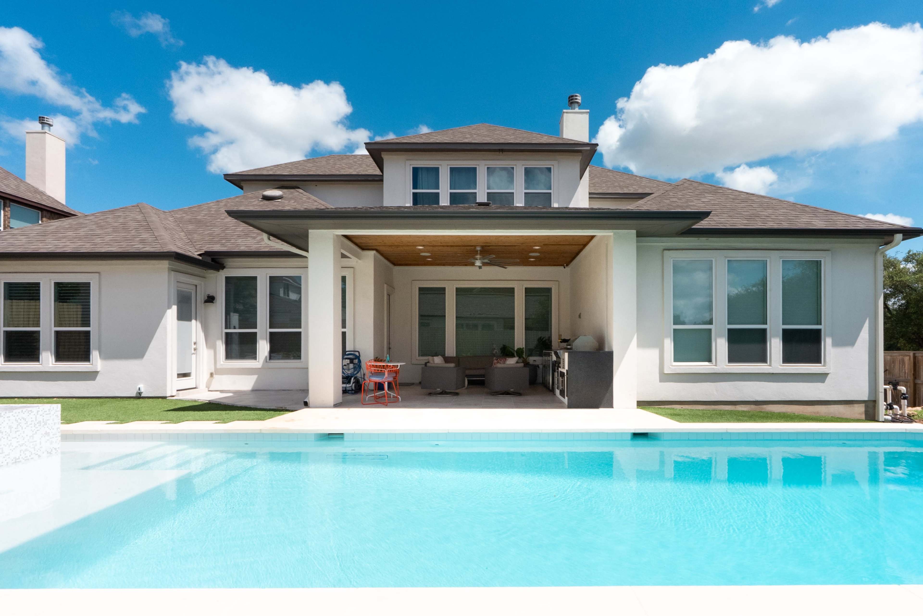 A modern house features a large, clear pool in the foreground and a spacious patio with multiple windows in the background.