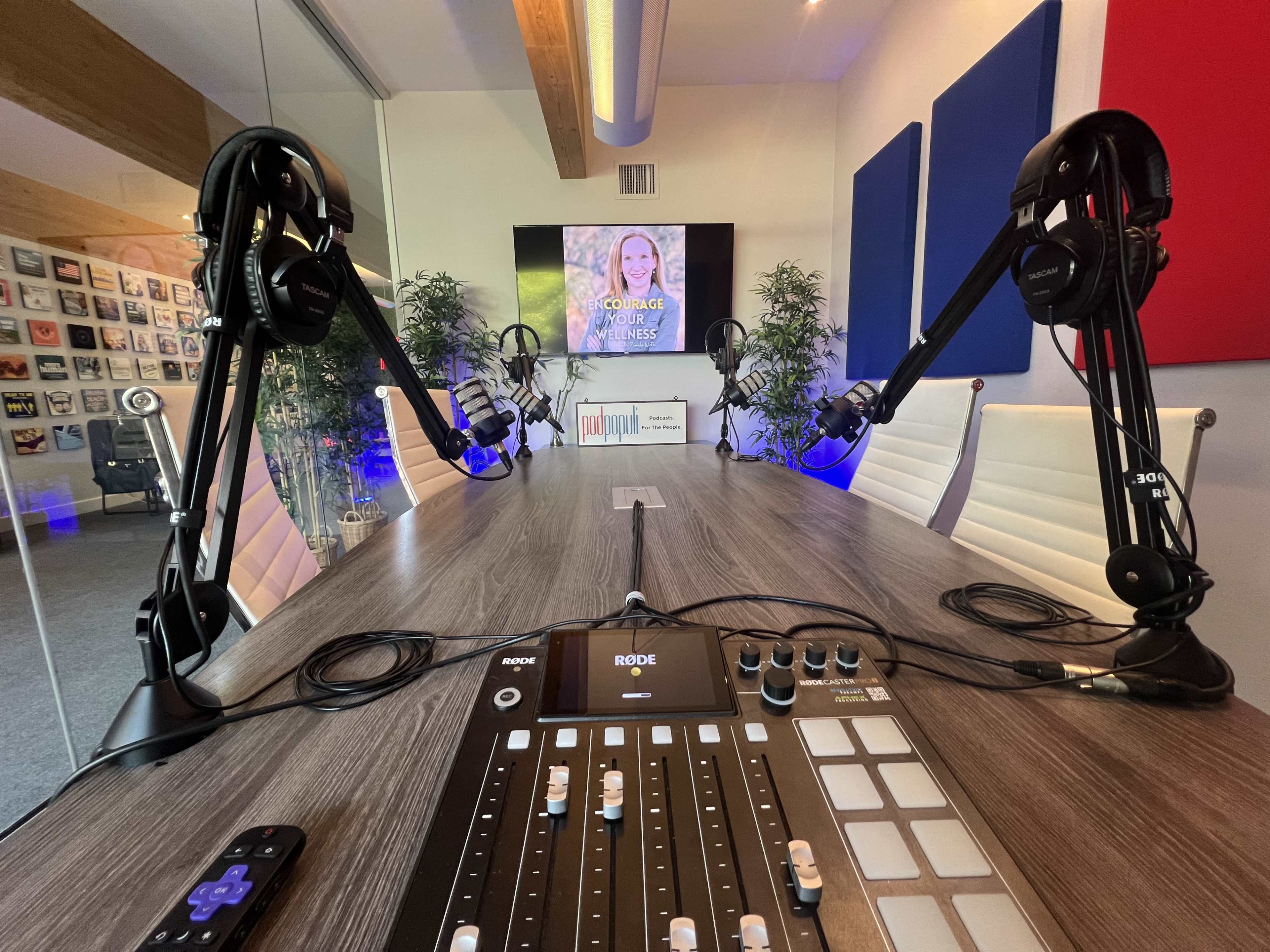 The image shows a modern recording studio with two microphone setups, a control panel, and a screen displaying a speaker's name in the background.