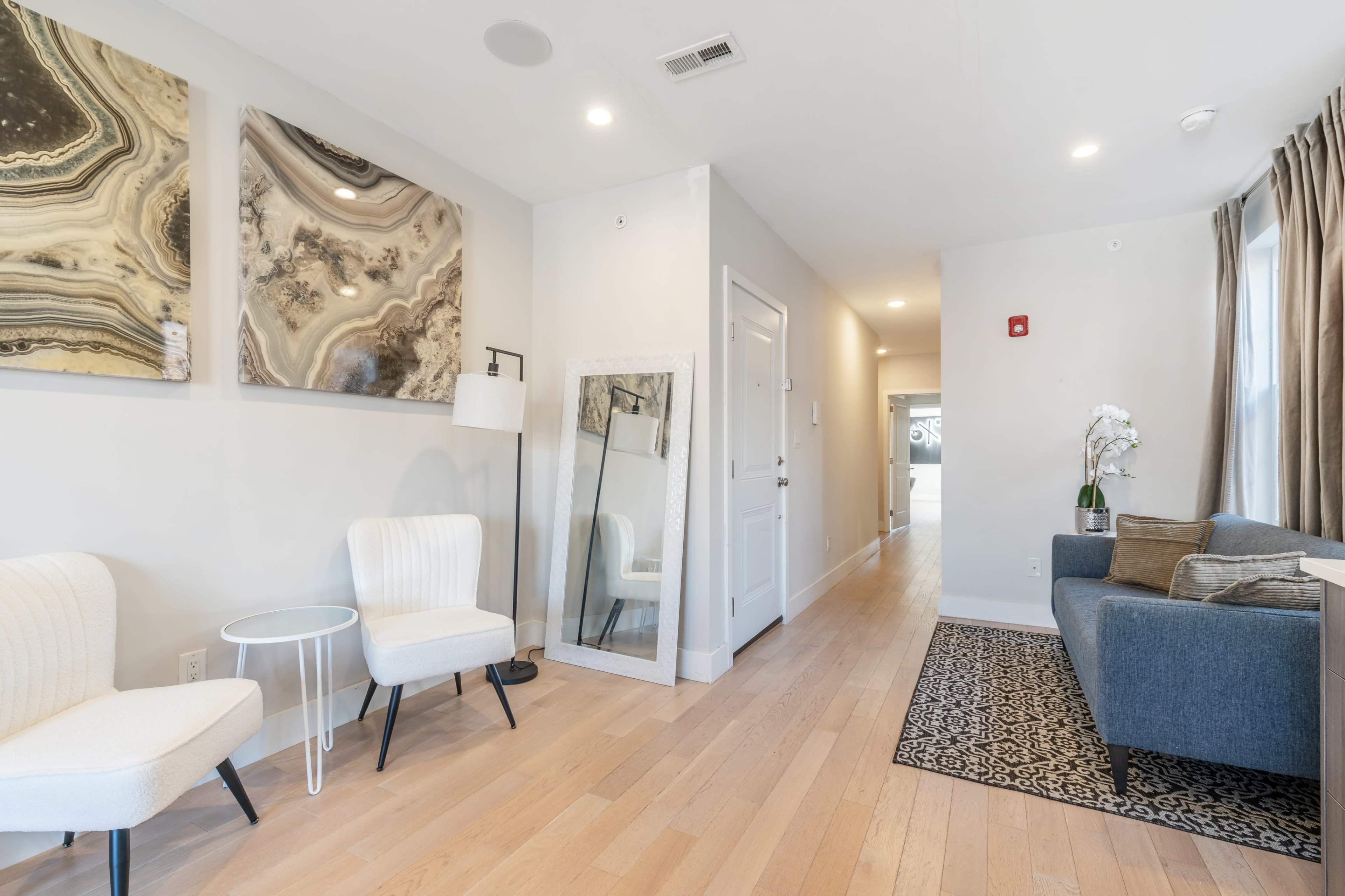 A modern living space with two white chairs, a full-length mirror, a gray sofa, and decorative wall art, leading to a hallway with a light-colored floor.