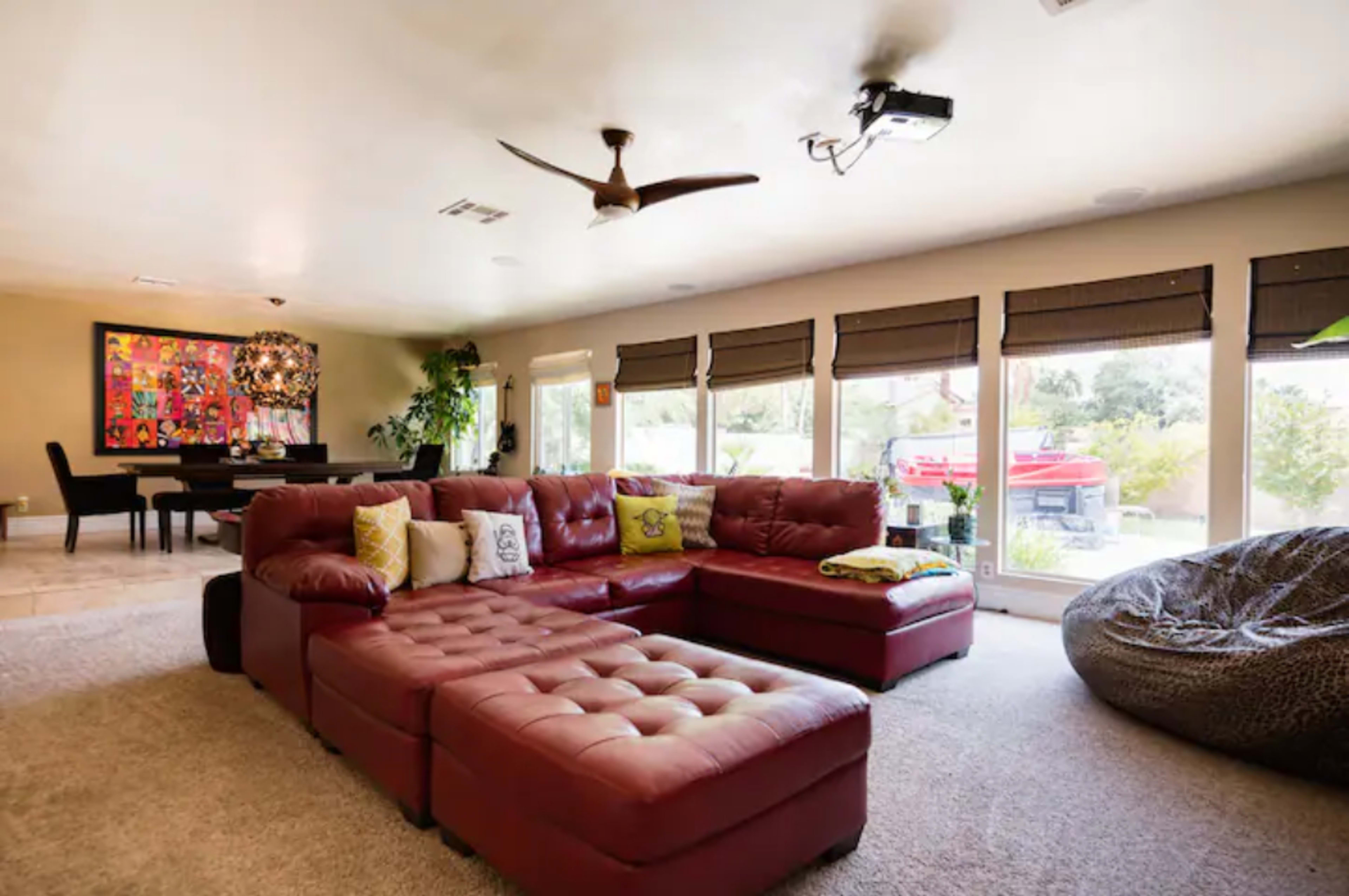 A spacious living room features a large red sectional sofa, a round ceiling fan, and multiple windows overlooking a garden.