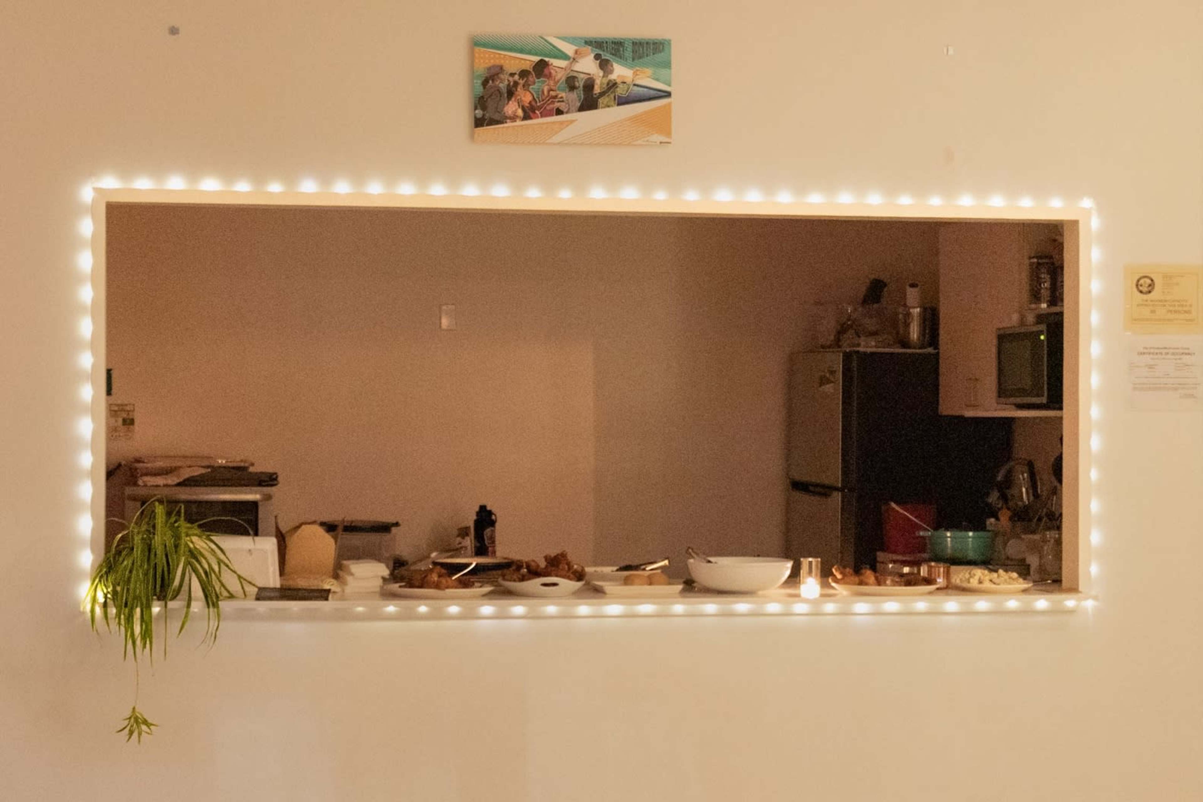 The image shows a kitchen area viewed through a well-lit serving window, with food items and kitchen appliances visible in the background.