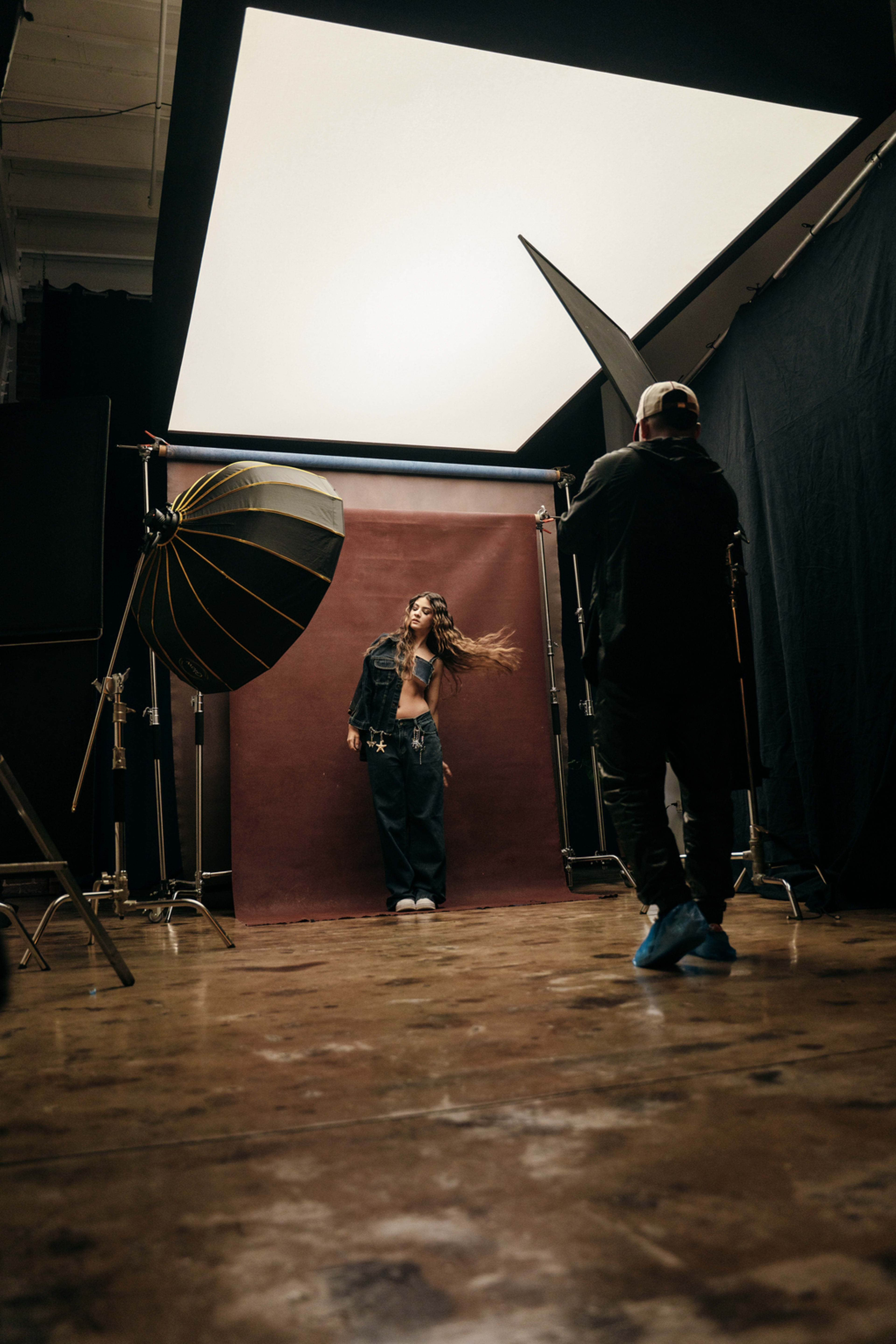 A person poses against a dark backdrop in a photography studio, while a photographer captures the shot using a large light source.