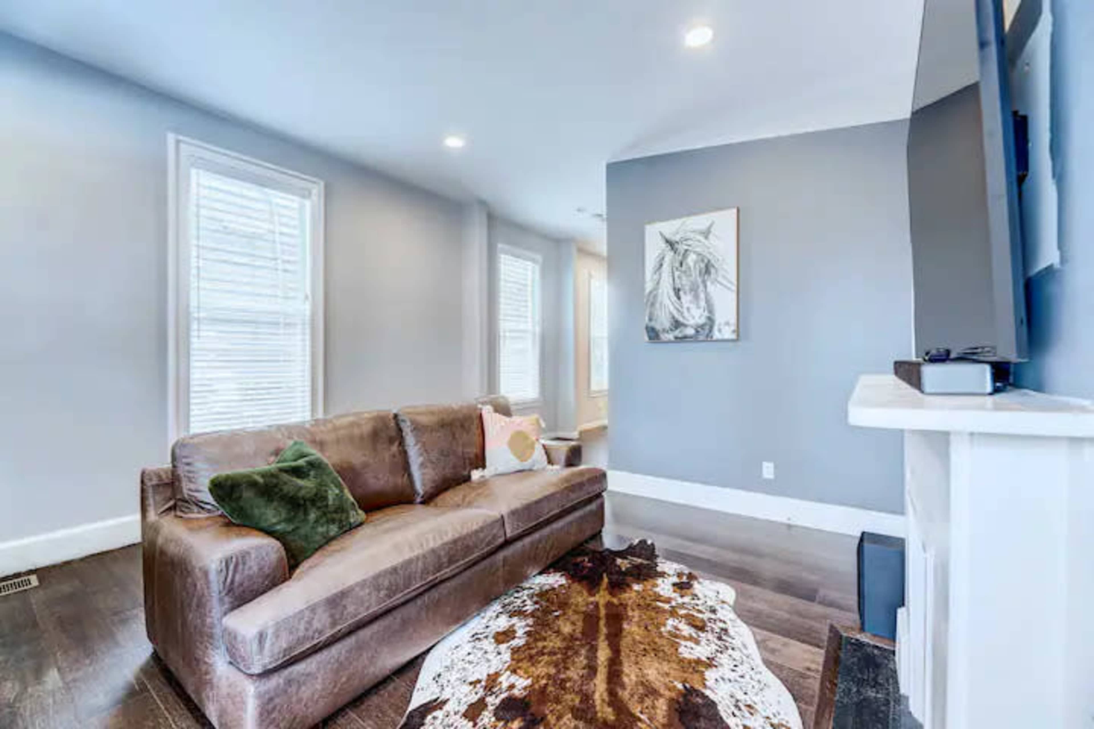 A living room features a brown sofa with a green pillow, a patterned rug, and a mounted television against a gray wall.