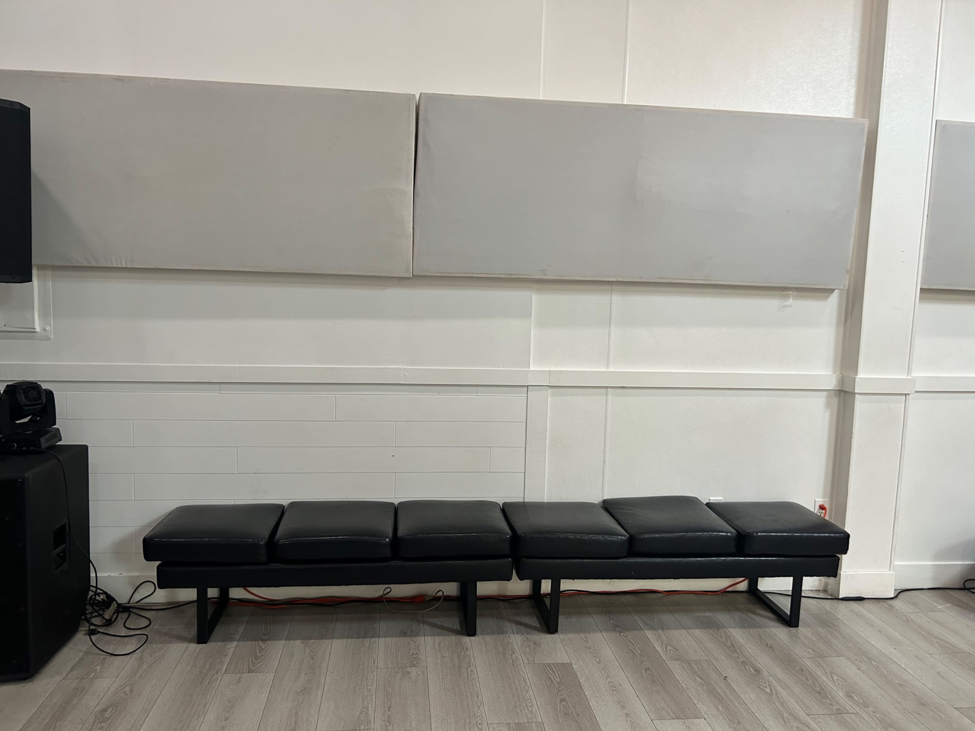 The image shows a long, black bench with multiple cushions positioned against a white wall, next to soundproofing panels.