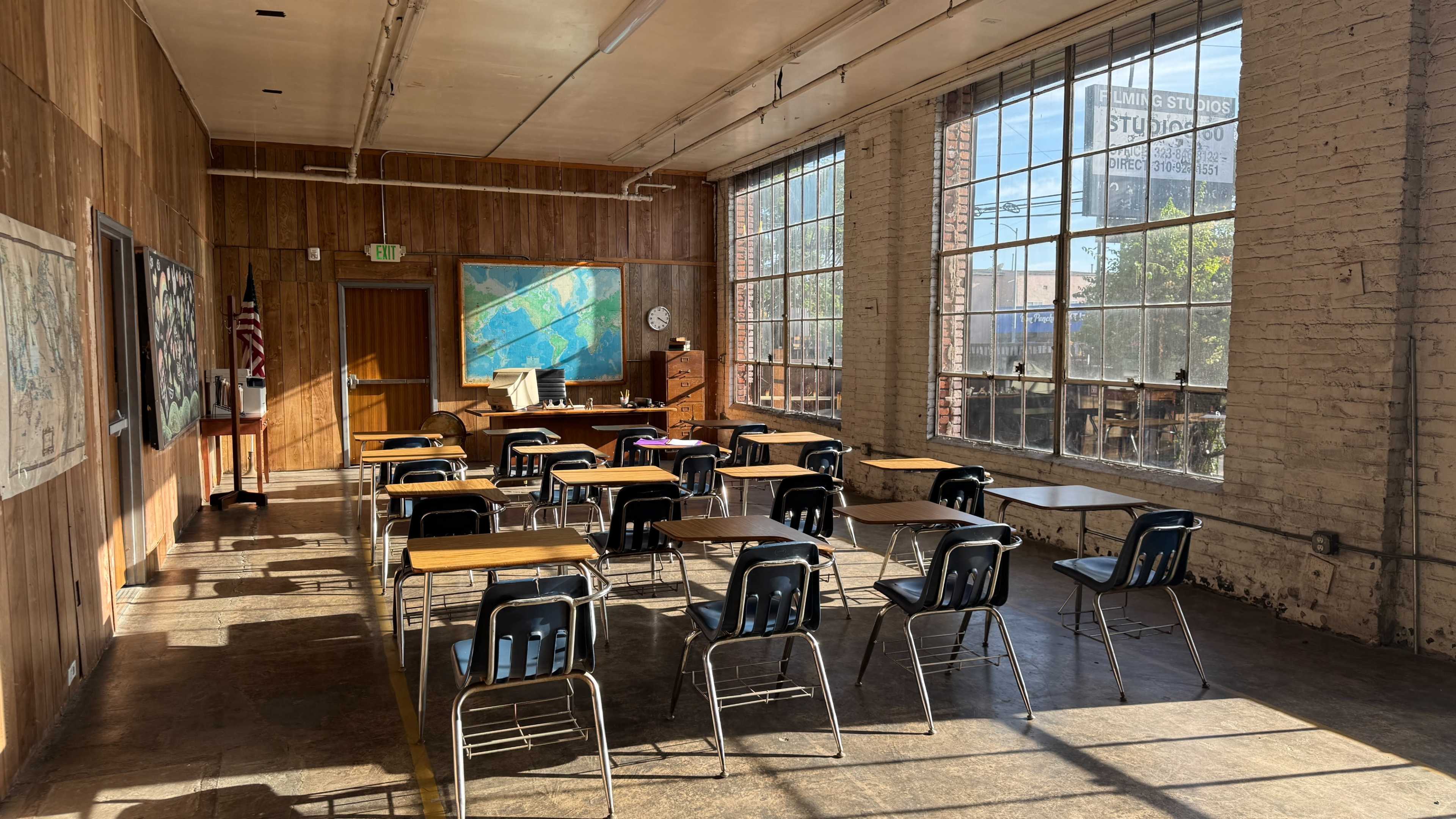 The image shows a classroom with wooden walls, several empty desks arranged in rows, and large windows allowing sunlight to enter.
