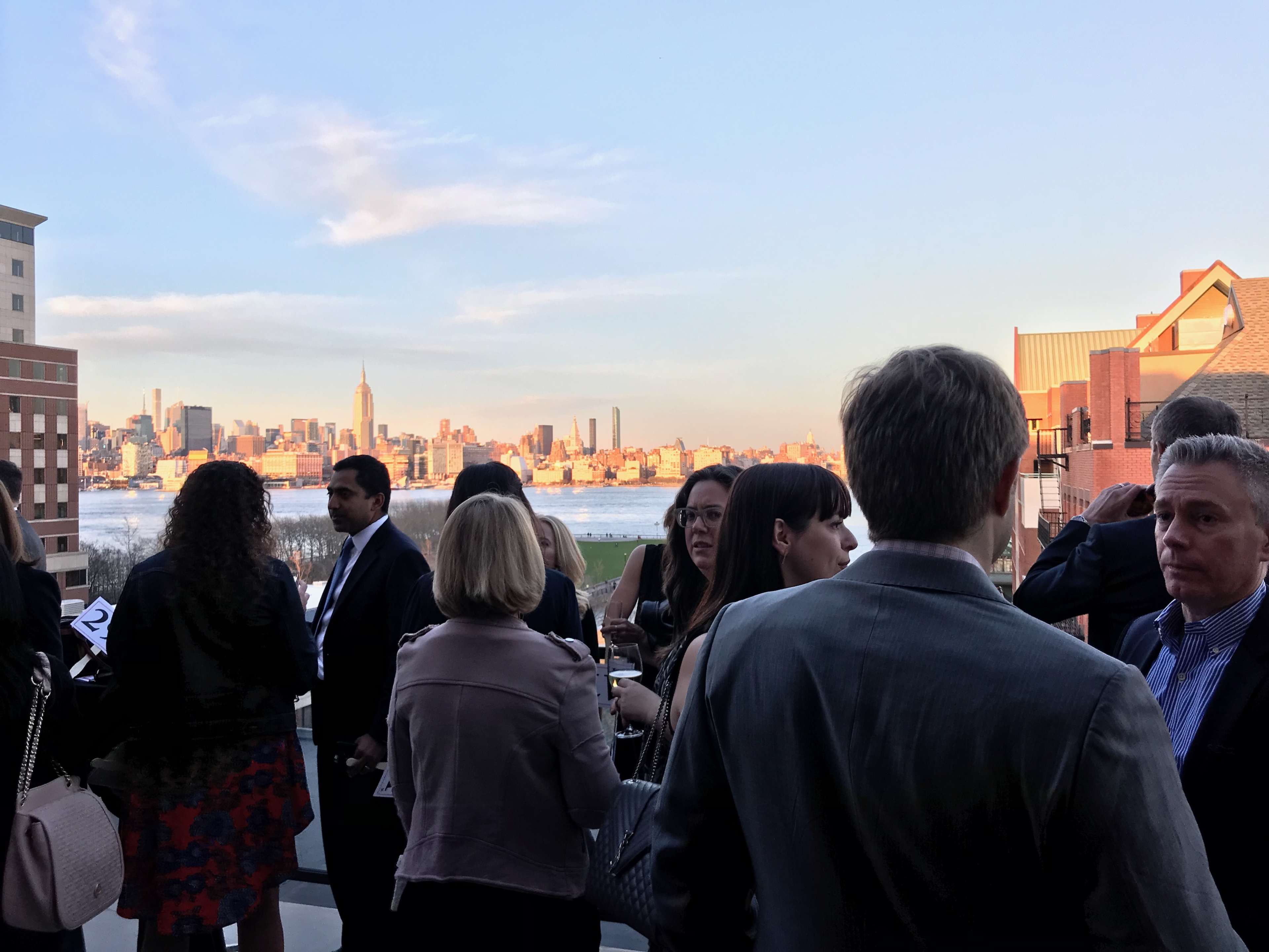 A group of people engages in conversation on a balcony overlooking the New York City skyline during sunset.