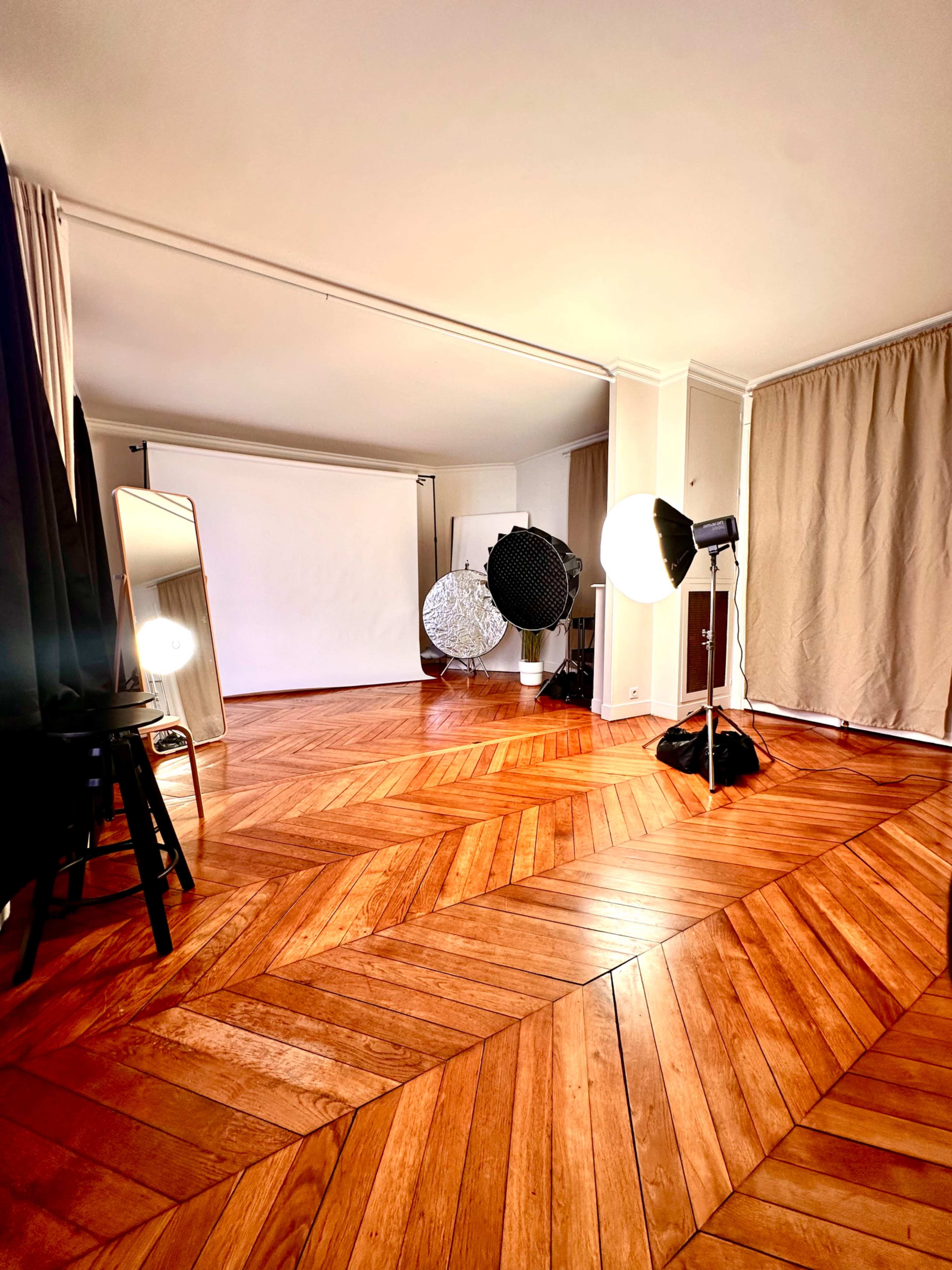 The image shows a studio interior featuring a wooden parquet floor, a white backdrop, and various lighting equipment arranged around the space.
