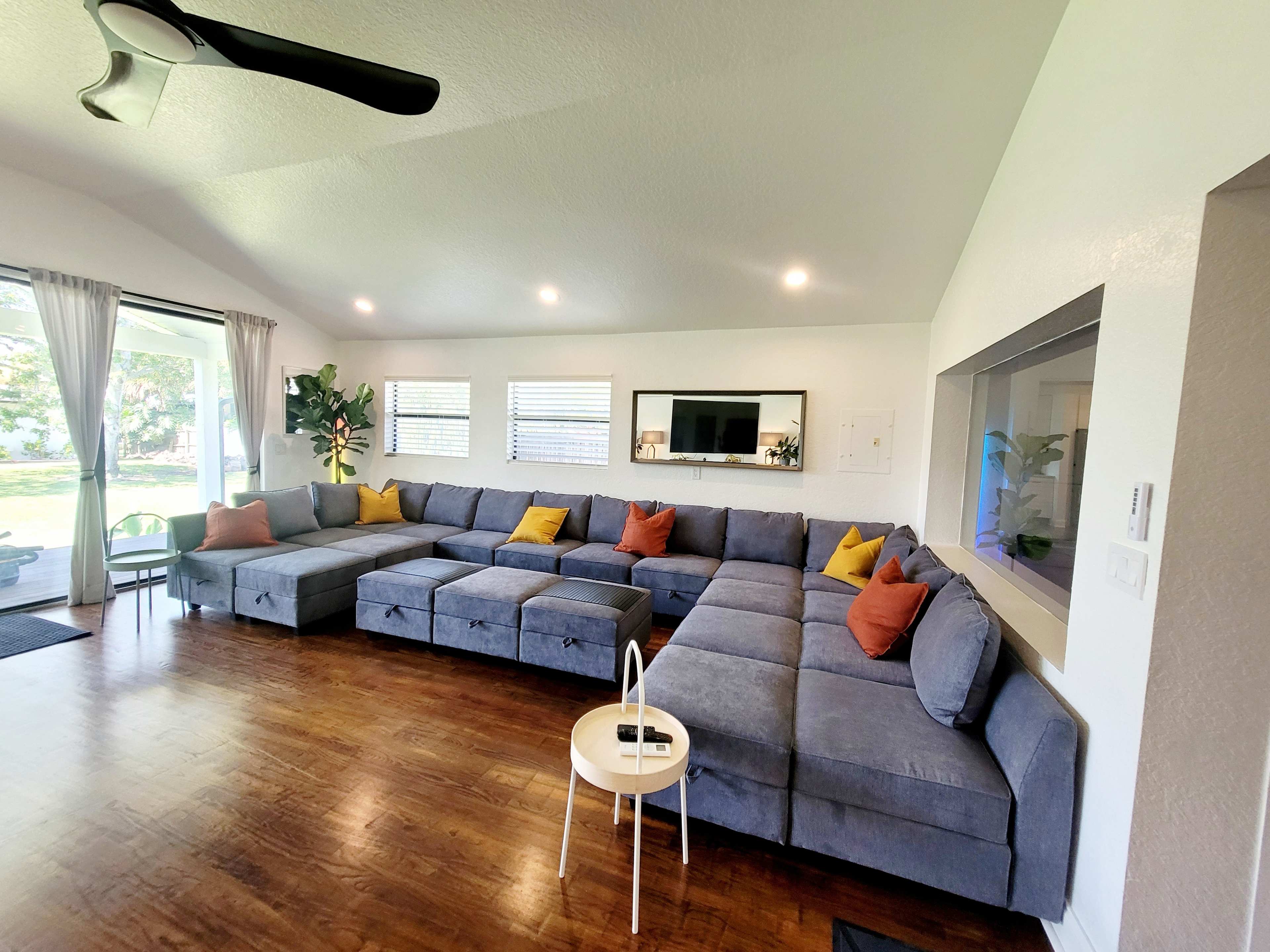 The image shows a spacious living room with a large, L-shaped sectional sofa covered in gray fabric, accented by colorful throw pillows, and a small white coffee table in front.