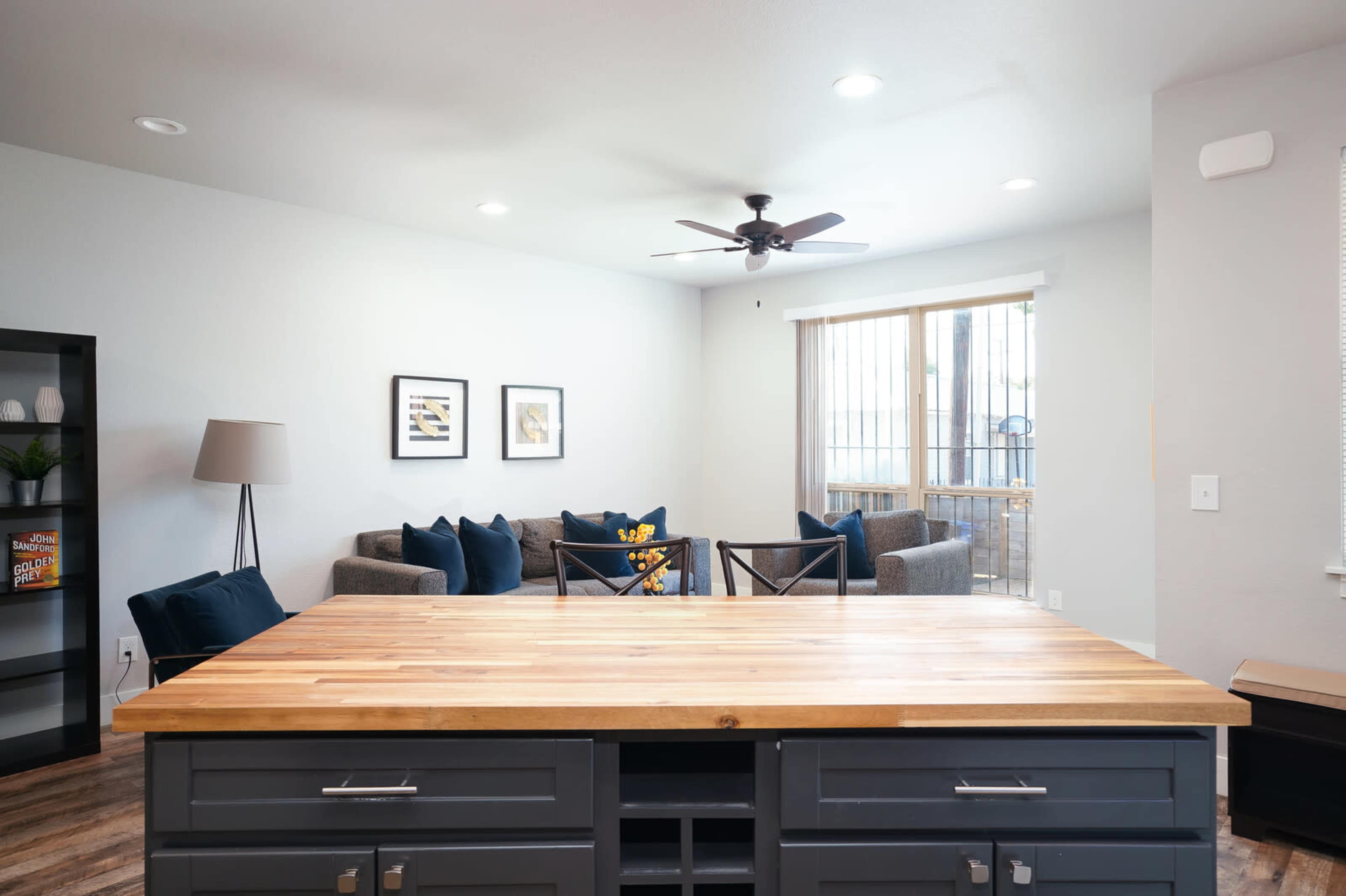A modern living space featuring a kitchen island with a wooden countertop in the foreground and a cozy seating area with gray couches and a ceiling fan in the background.
