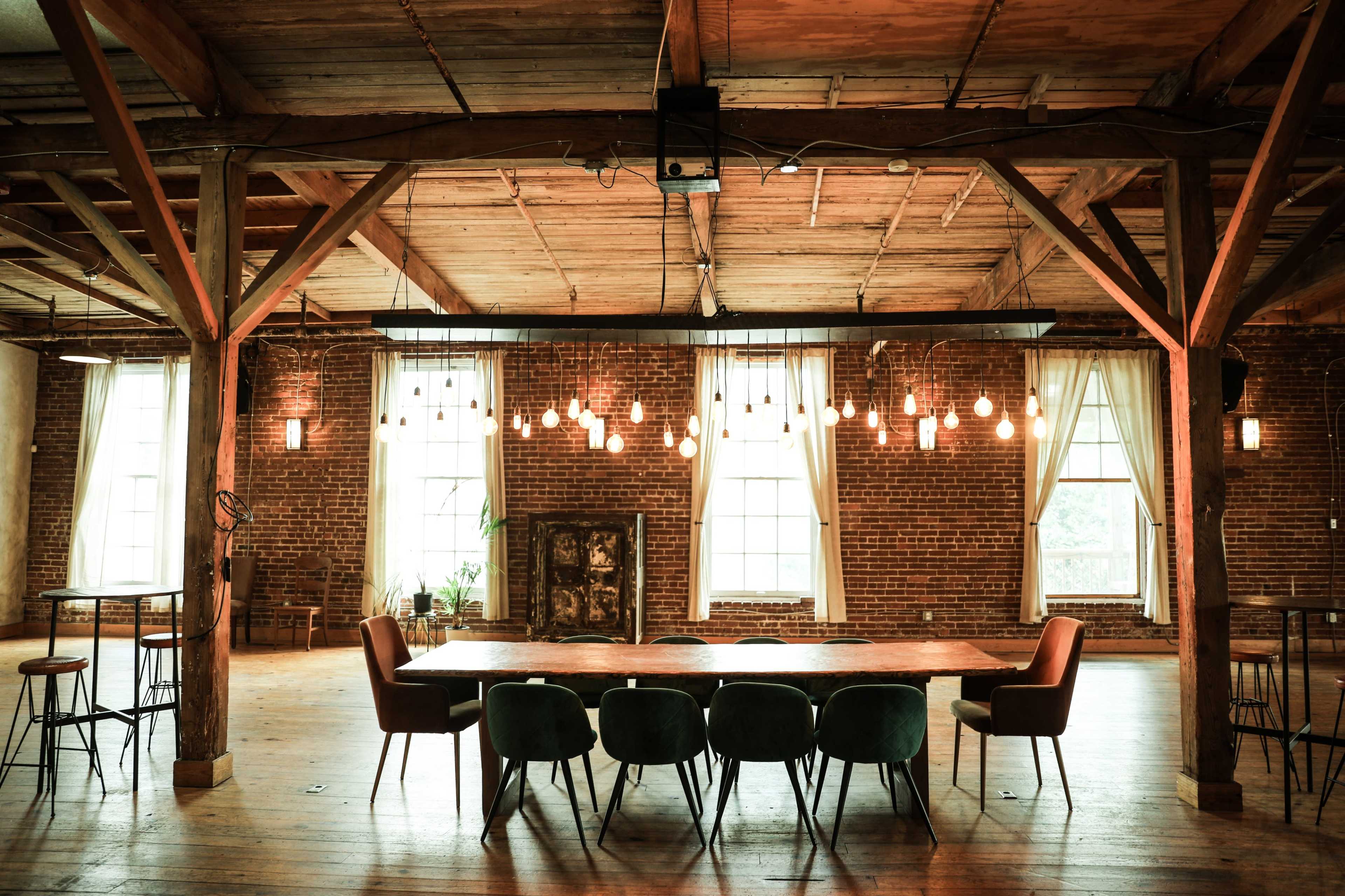 The image shows an industrial-style interior with wooden beams, exposed brick walls, a long table surrounded by chairs, and pendant lights hanging from the ceiling.
