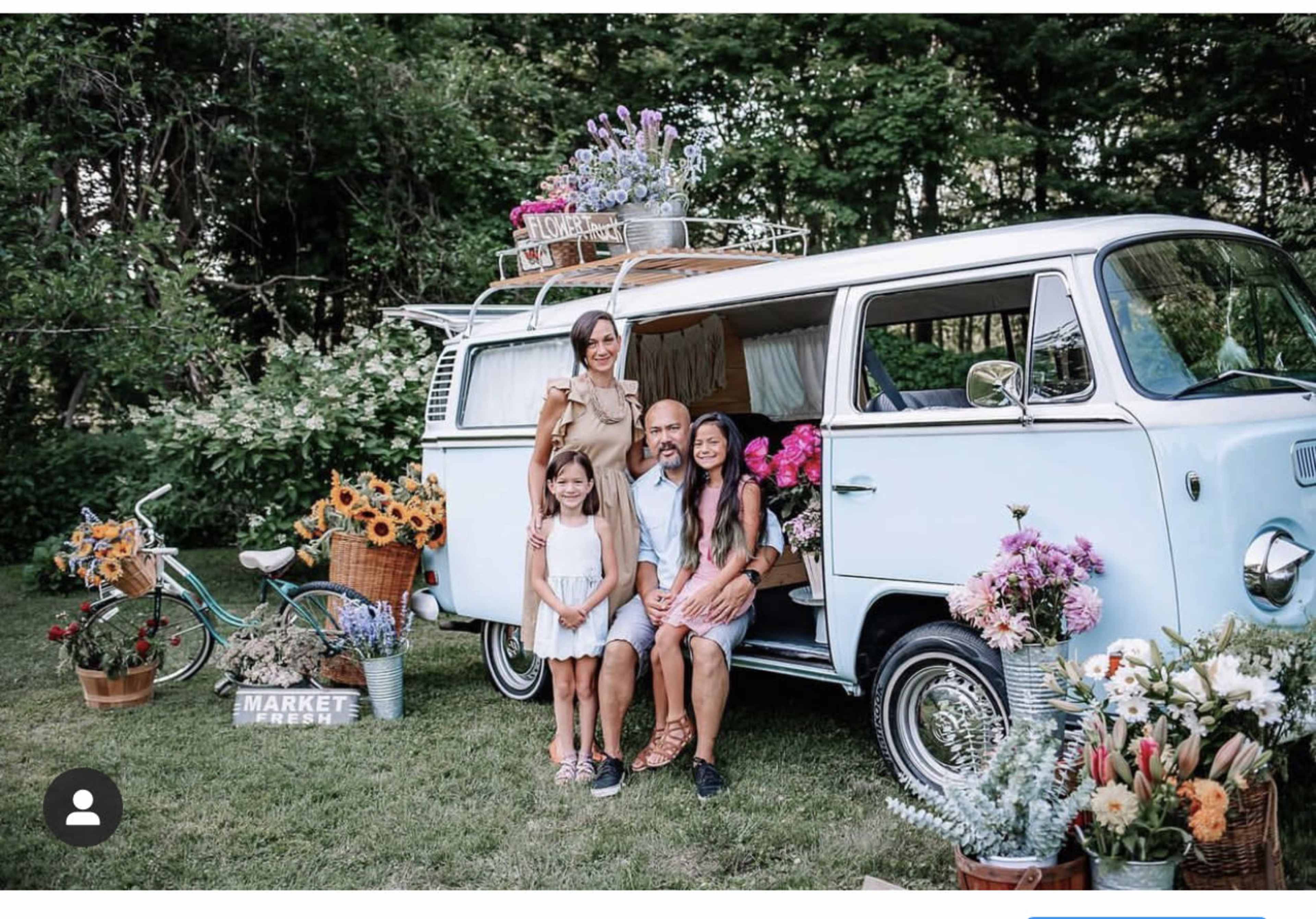 A family poses in front of a vintage blue van surrounded by colorful flower arrangements and decorative items in a garden setting.