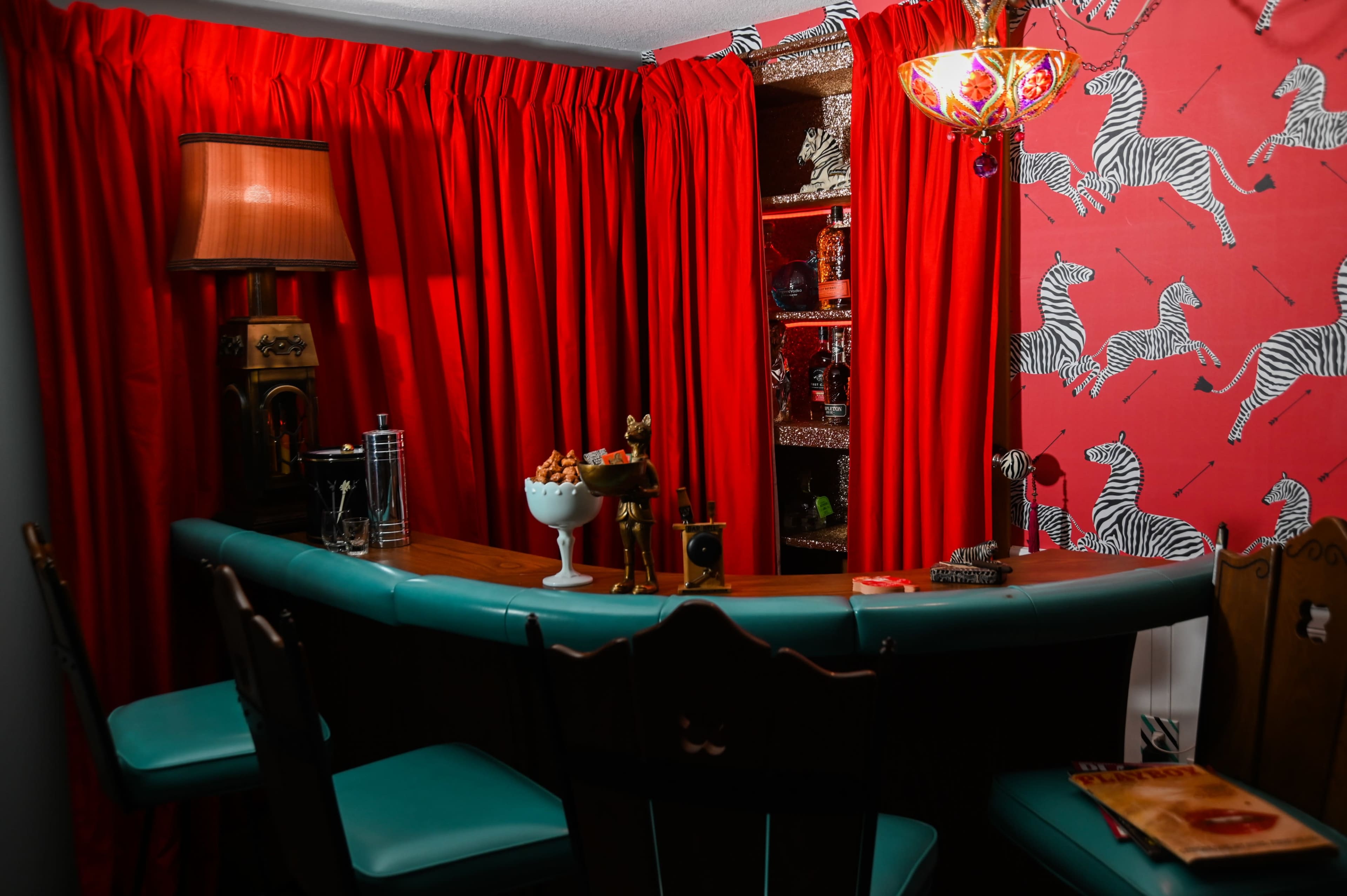 A home bar features a wooden counter with turquoise seating, surrounded by red curtains and zebra-patterned wallpaper.