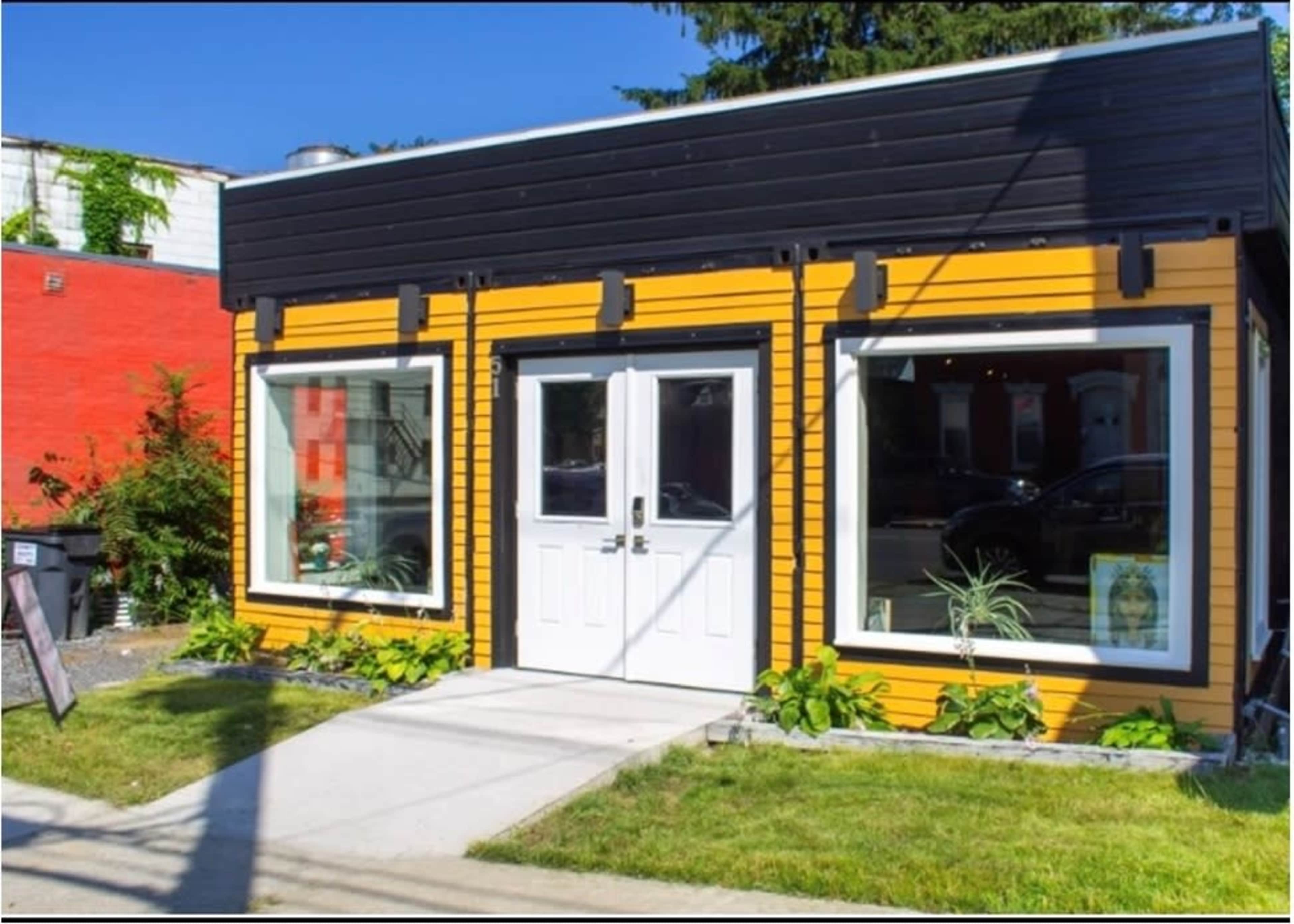 A small, brightly colored yellow building with large windows and a white door, situated next to a red structure.