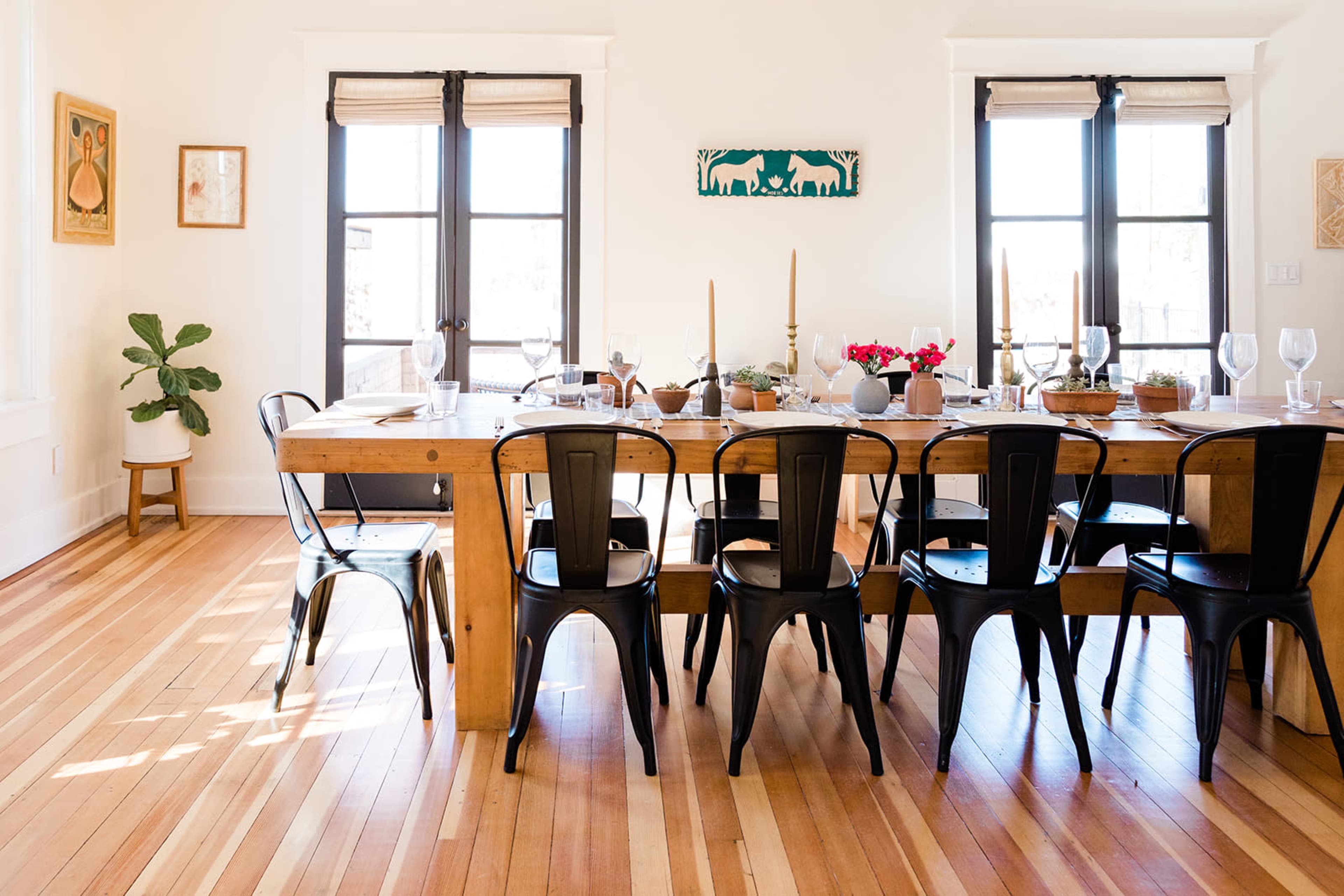 A wooden dining table is set with dishes and glassware in a bright room featuring large windows and a plant in the corner.