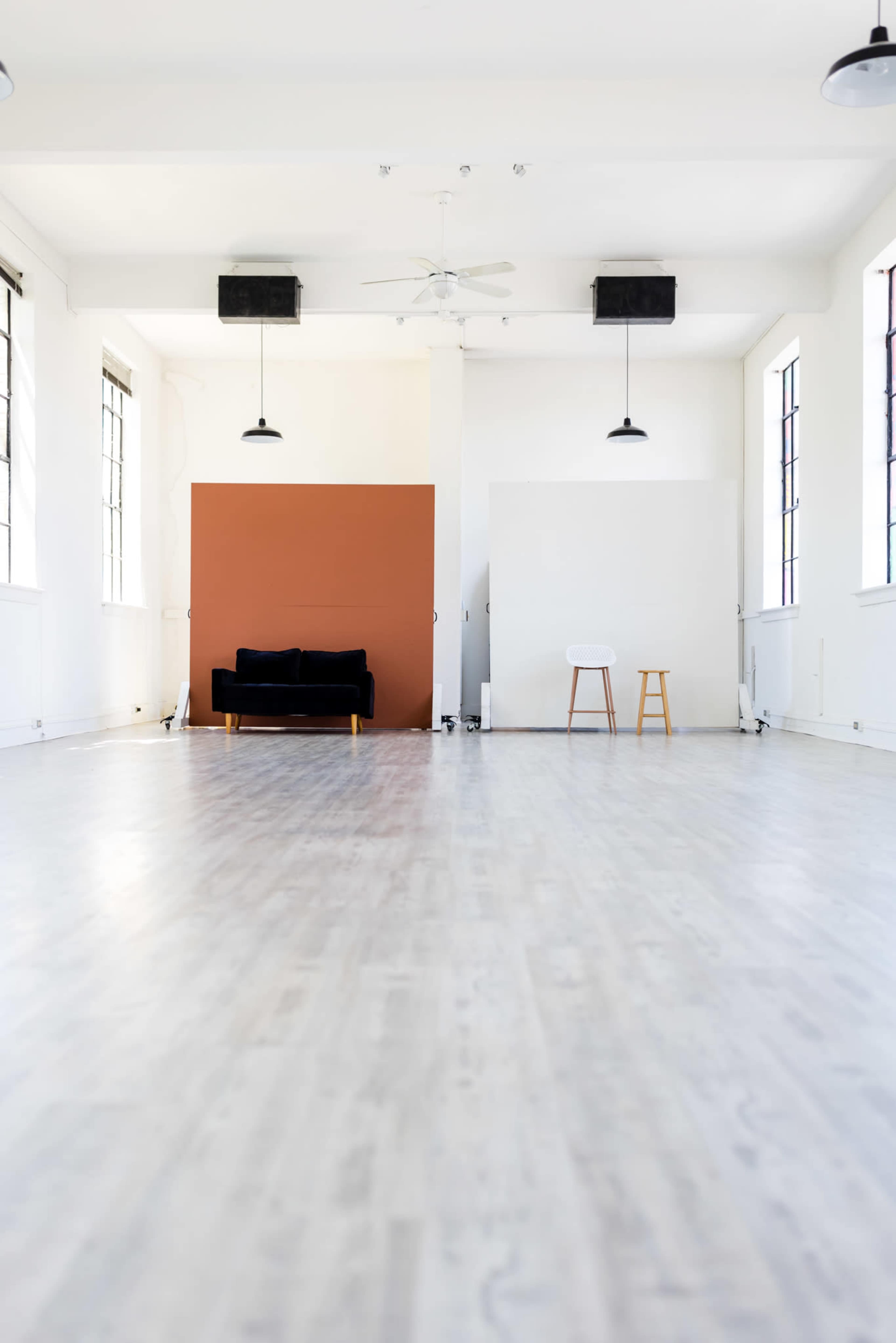 A spacious, minimalist room with wooden flooring, featuring a black sofa against a brown accent wall and a white wall opposite it, along with two pendant lights.