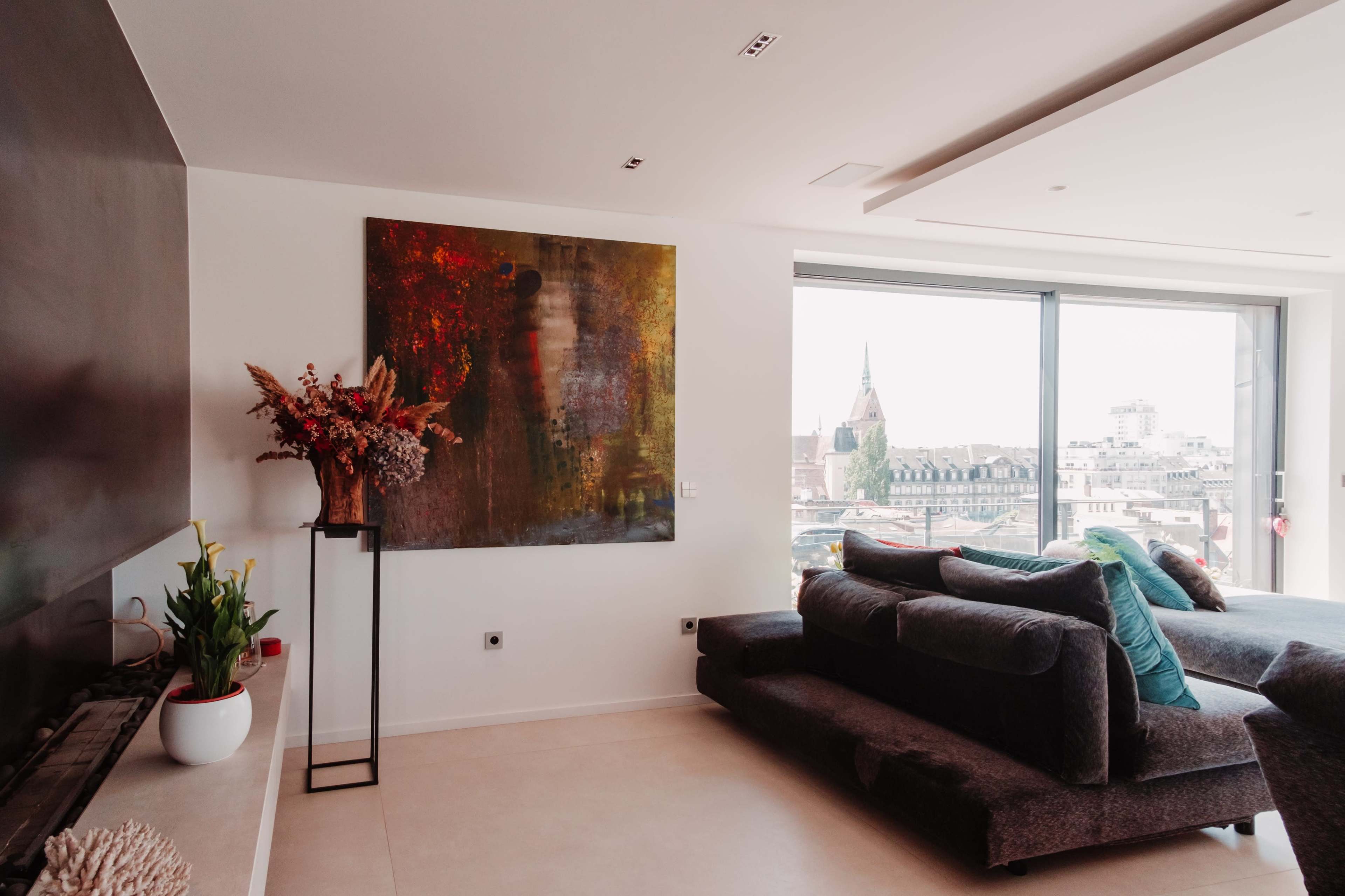 A modern living room features a dark sofa, a large abstract painting, and a window overlooking a cityscape.