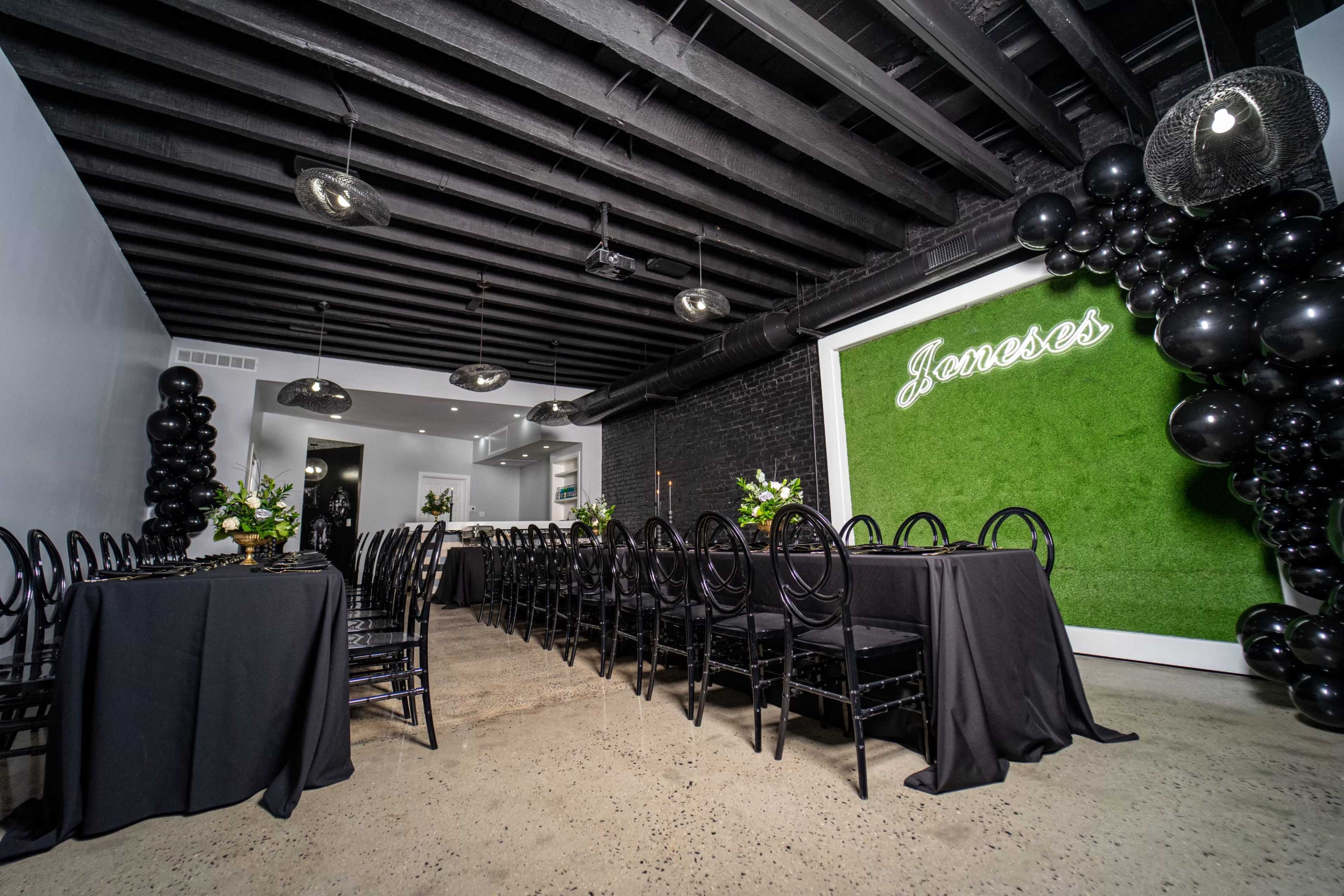 A spacious event room with black tables, chairs, and decor, featuring a green grass wall with "Javoses" in illuminated letters.