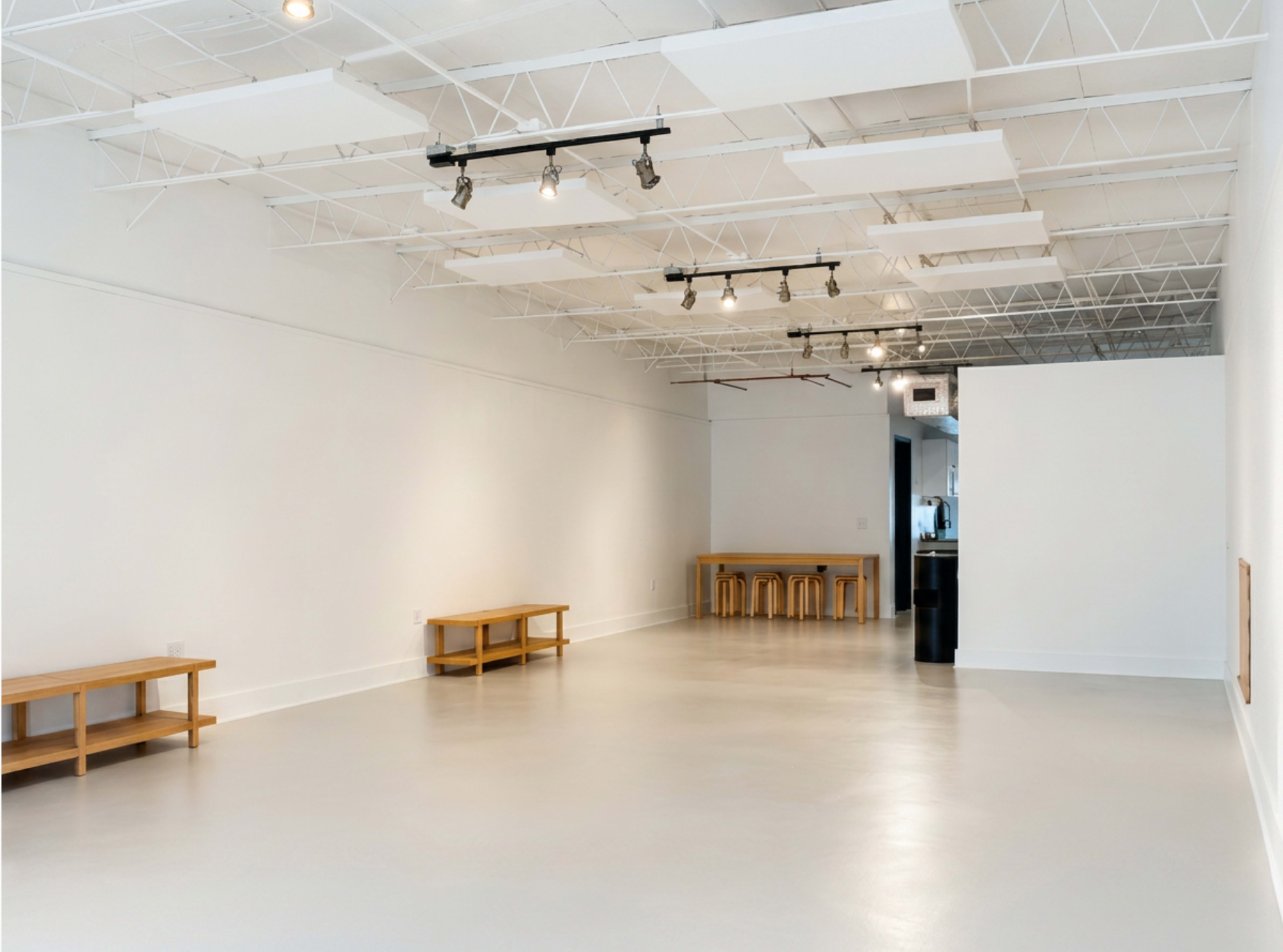 The image shows a spacious, empty gallery with white walls, a polished concrete floor, and wooden furniture along the sides.