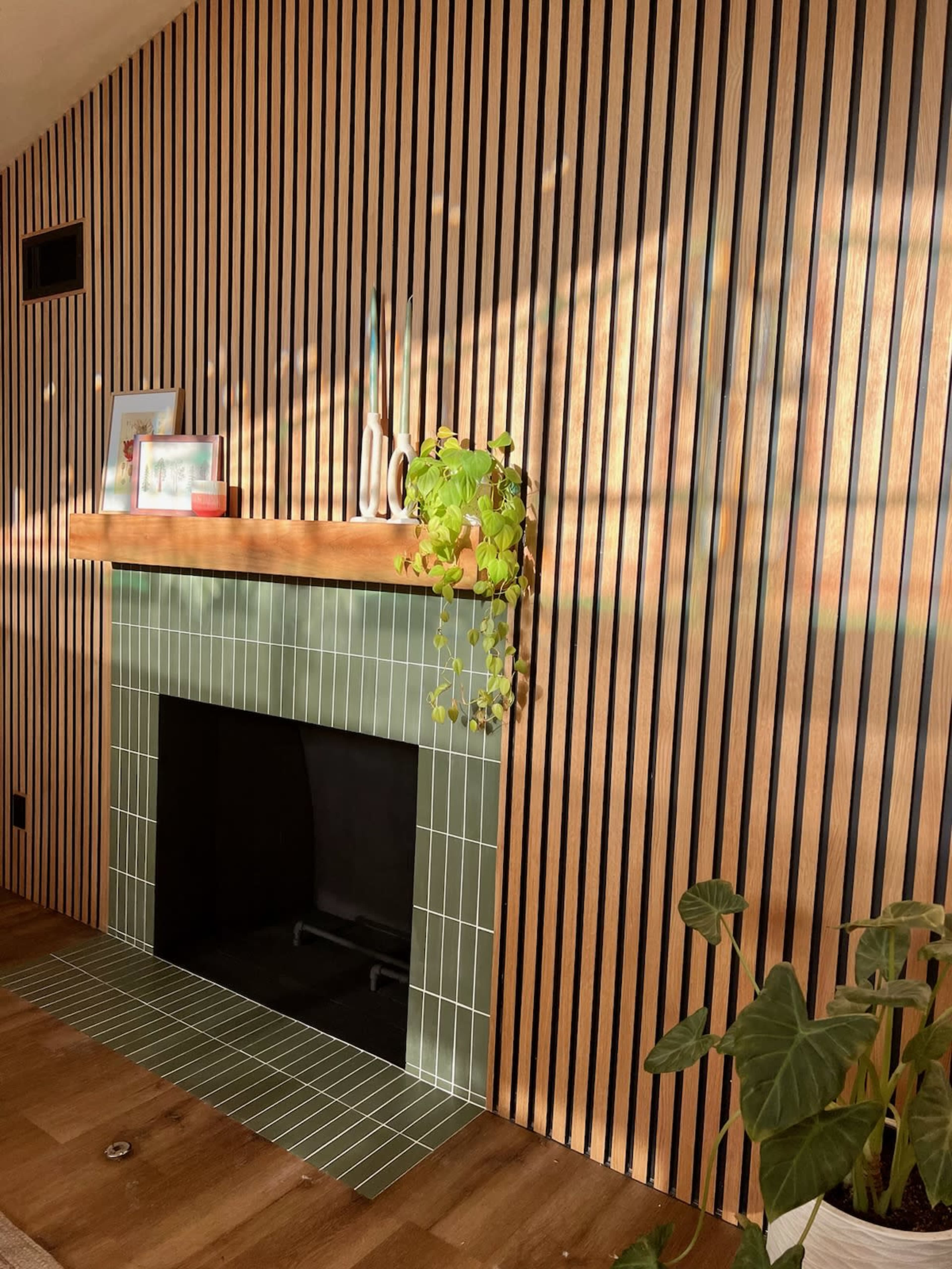 A modern interior featuring a green tiled fireplace with a wooden mantel and a striped wood wall, along with a potted plant in the foreground.