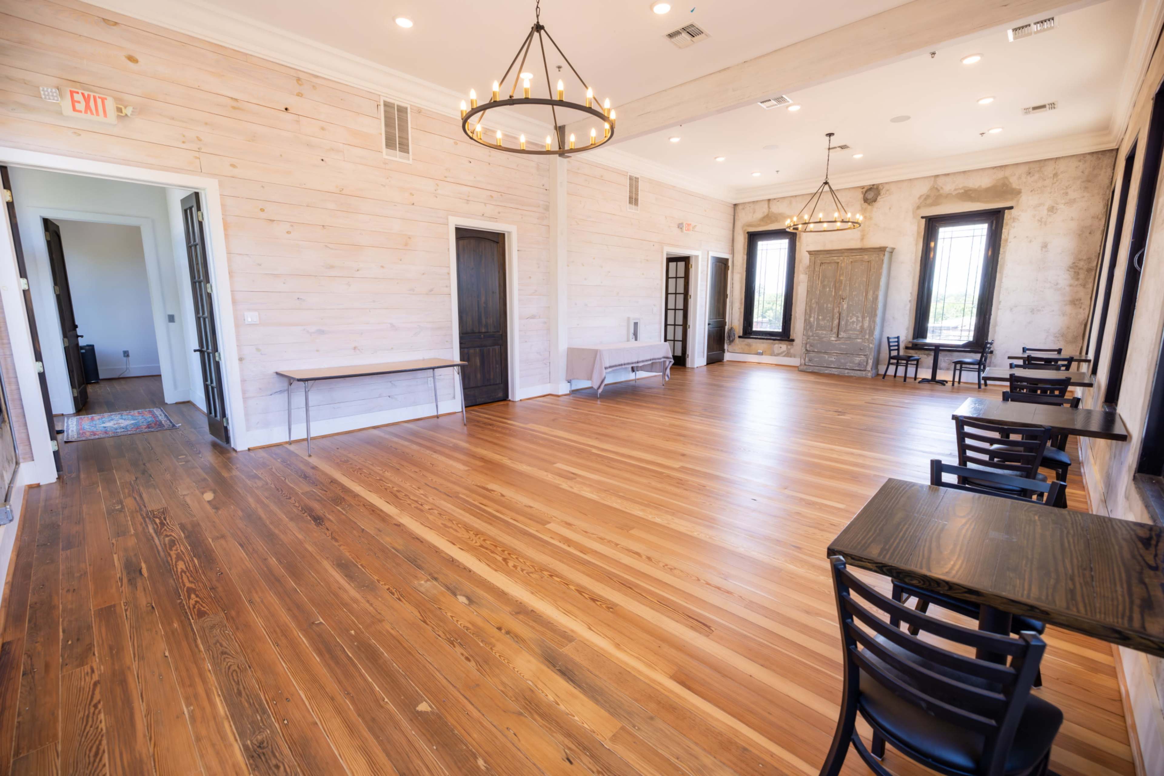 The image depicts a spacious interior room featuring wooden floors, light-colored wooden walls, and several tables with chairs, illuminated by hanging chandeliers.