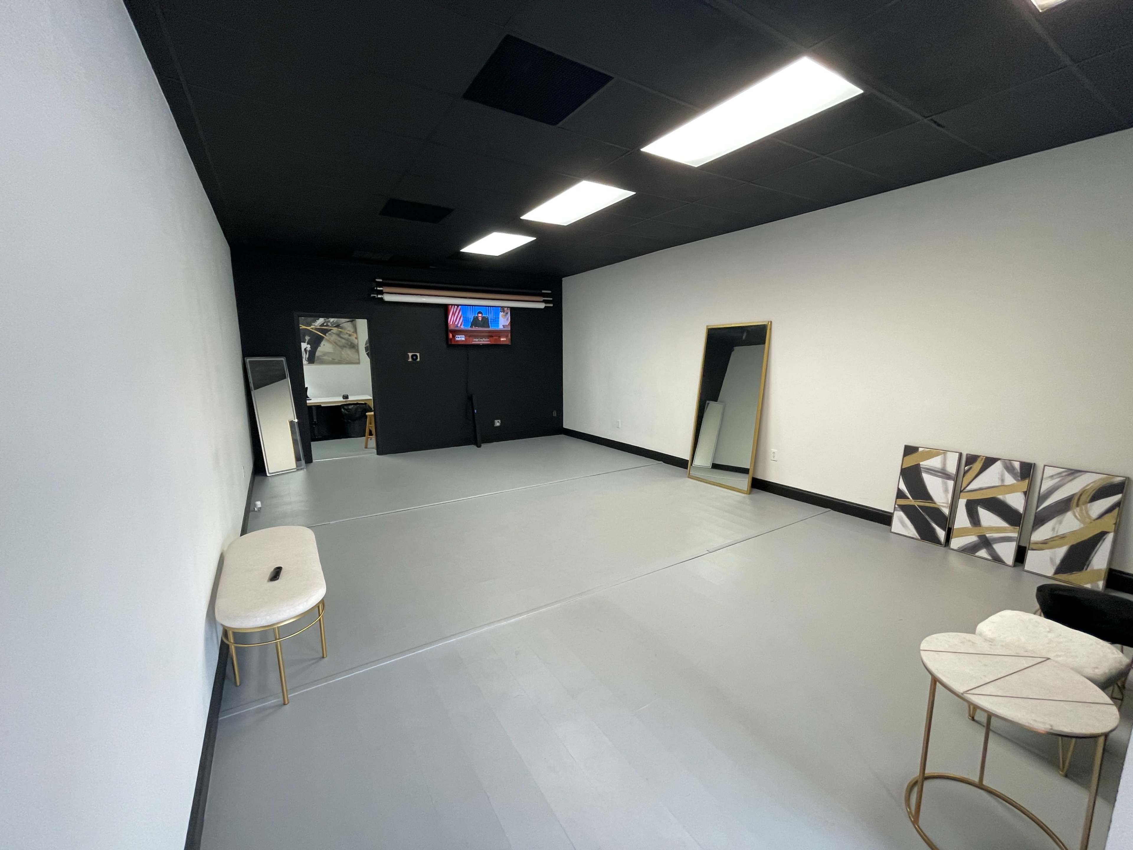 The image shows a sparse, empty room with gray flooring, a large mirror on one wall, a television mounted on another, and minimal furniture including a small bench and a couple of tables.