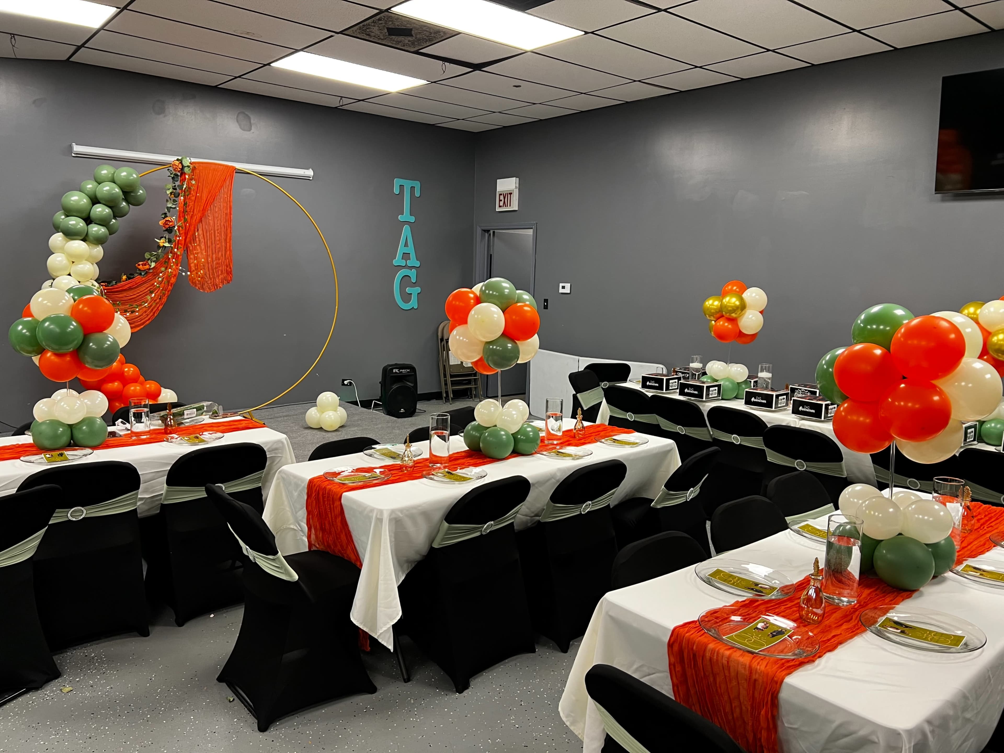 A decorated event space with tables set for a gathering, featuring orange and green table runners, balloon arrangements, and a backdrop with the word "TAG."