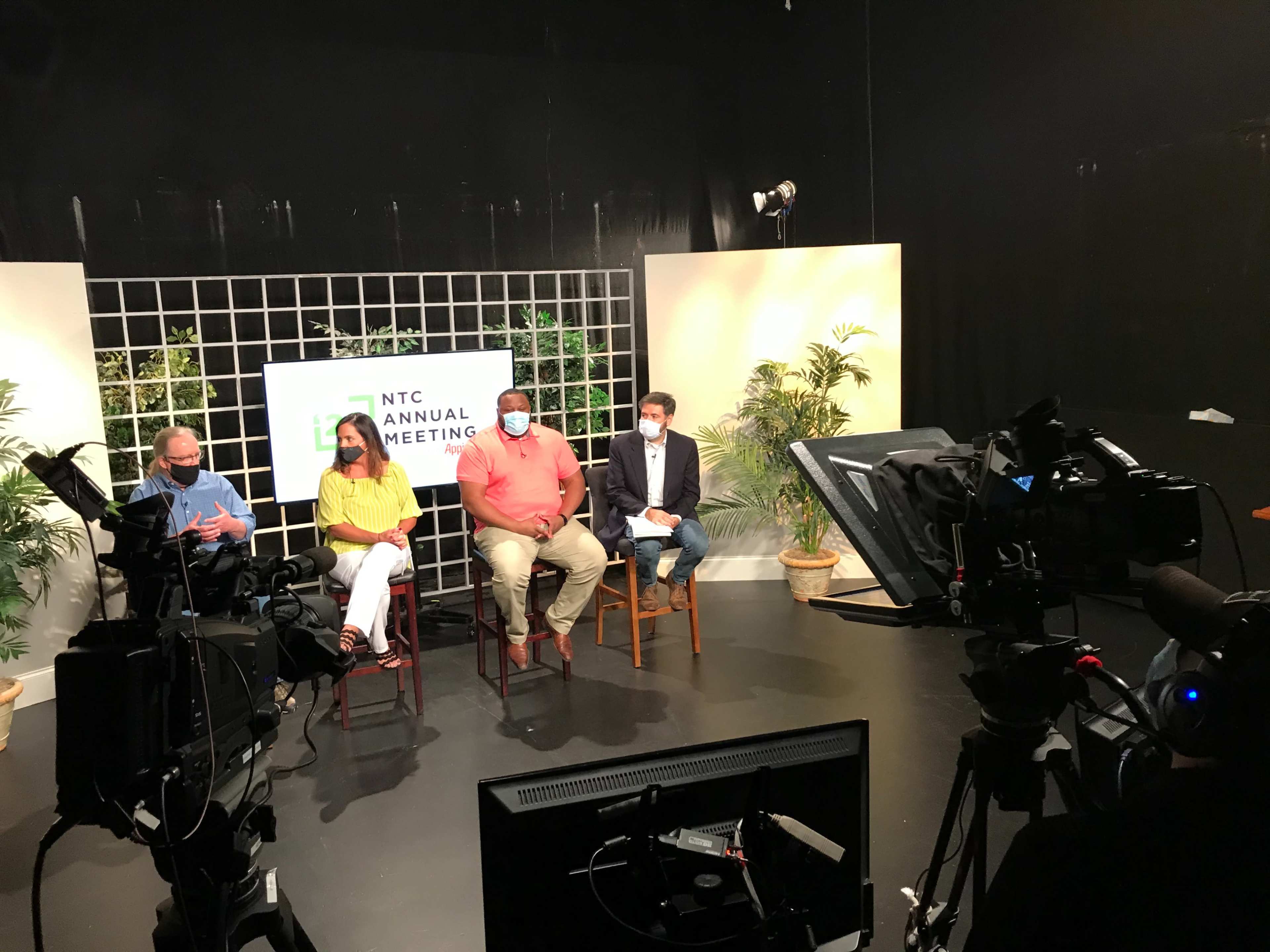 A group of four people are seated on stools in a studio setting for a meeting, with a sign reading "NTC Annual Meeting" in the background and cameras set up to record.
