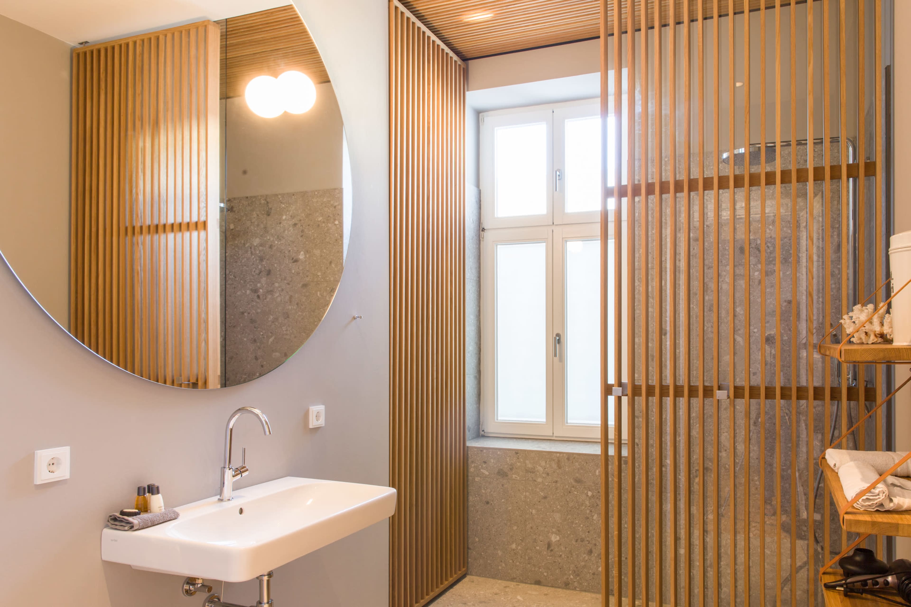 The image shows a modern bathroom with a round mirror, a wall-mounted sink, and wooden slatted partitions separating the shower area from the rest of the space.