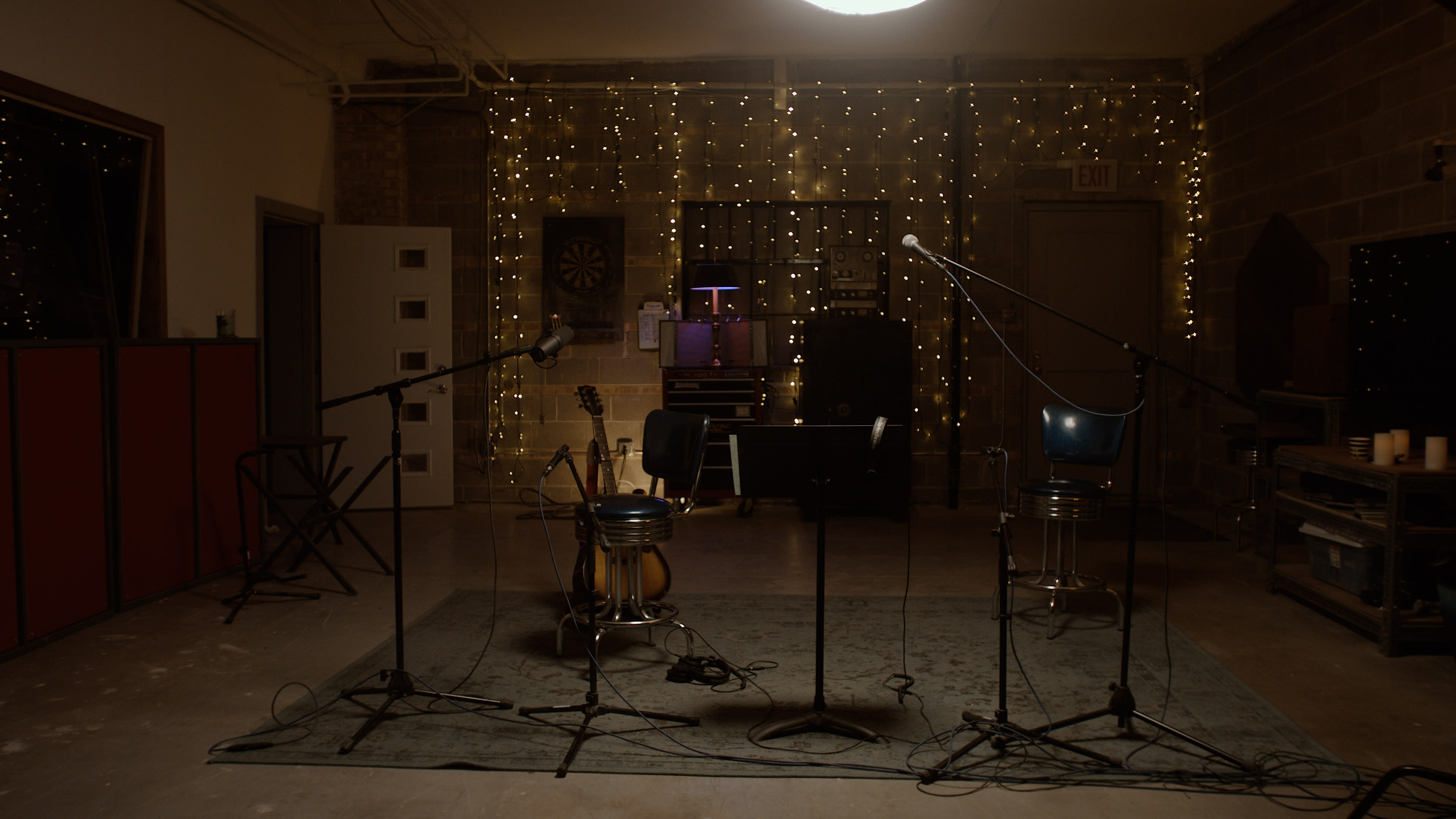 The image shows a recording studio with microphones set up on a rug, an acoustic guitar leaning against a chair, and string lights illuminating the background.