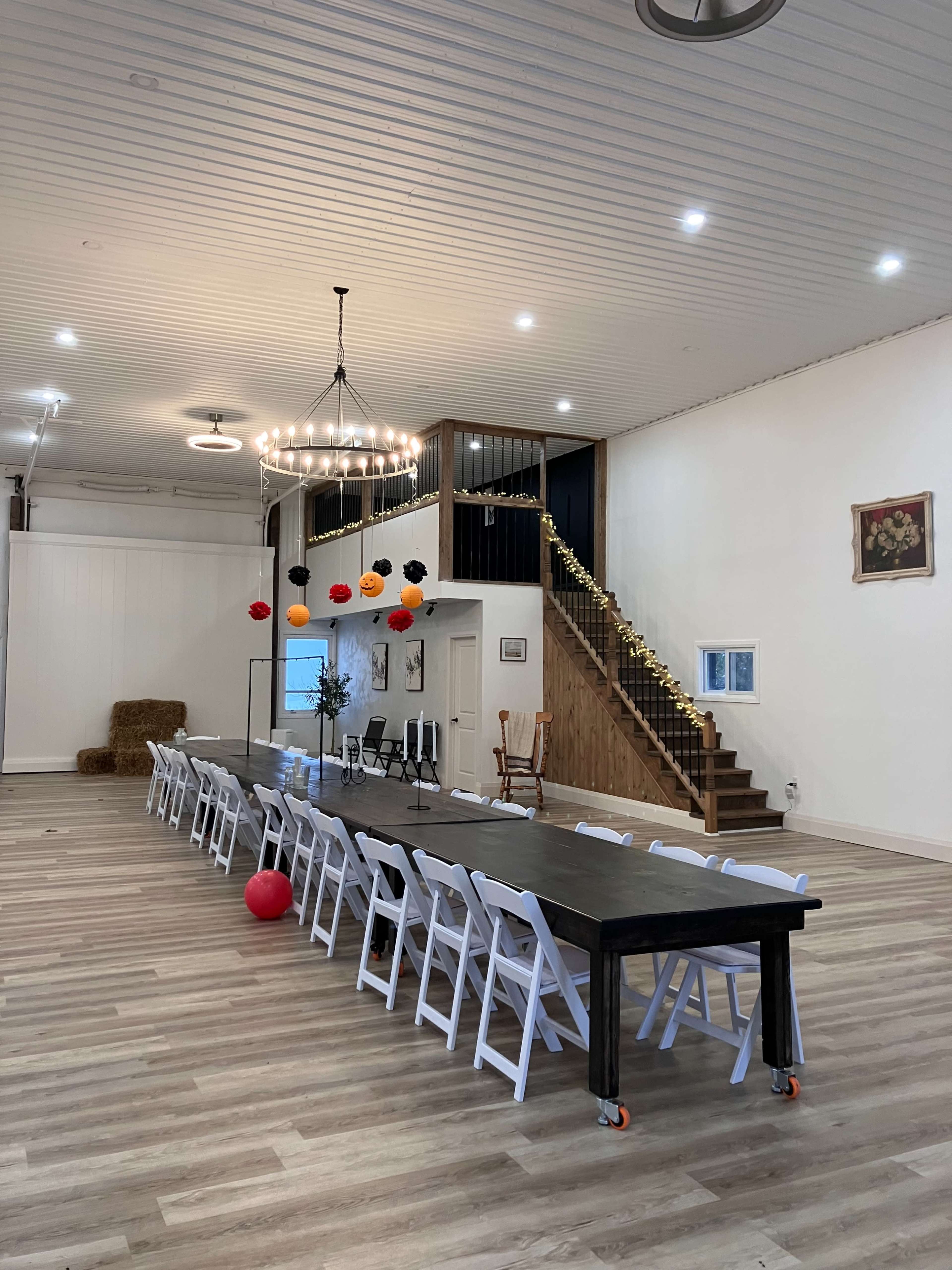 The image shows a spacious event hall with a long black table surrounded by white chairs, leading to a staircase decorated with orange and black balloons.