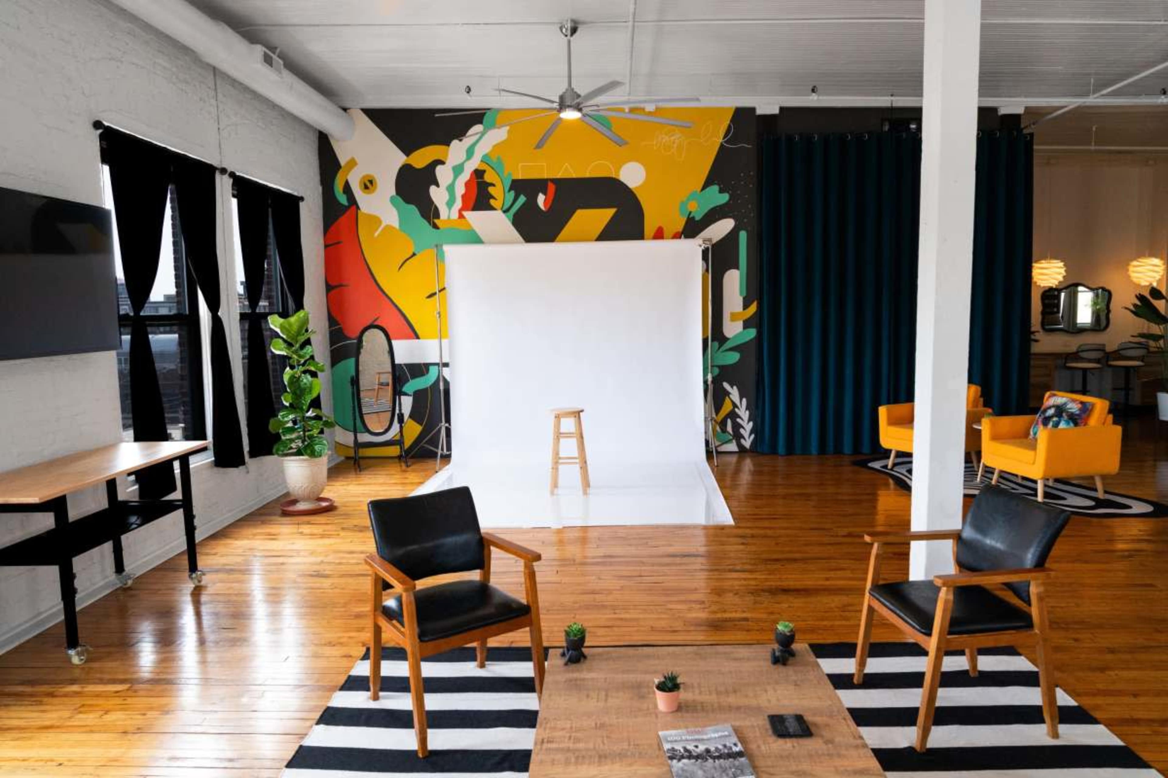 The image shows a spacious interior with a white backdrop, wooden flooring, plants, colorful seating areas, and a modern mural on the wall.